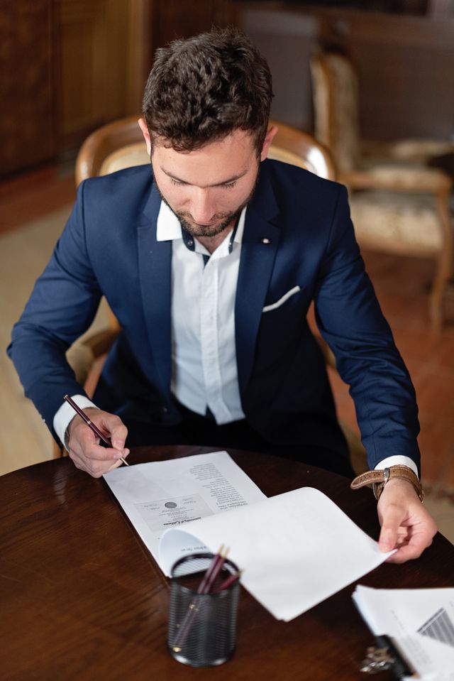Homme en costume bleu signant un document à une table en bois dans un intérieur classique