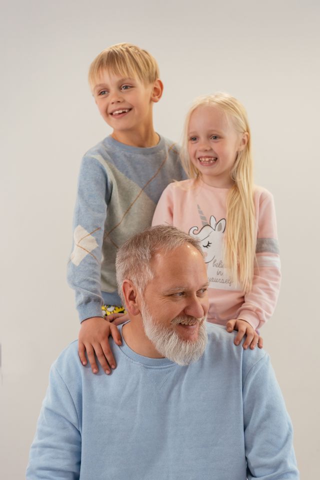 Grand-père barbu assis, ses deux petits-enfants souriants debout derrière lui, sur fond clair