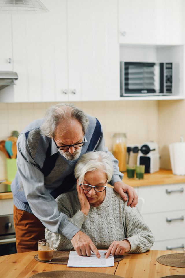 Couple de retraités consultant ensemble un document à la table de leur cuisine, l'un debout derrière l'autre