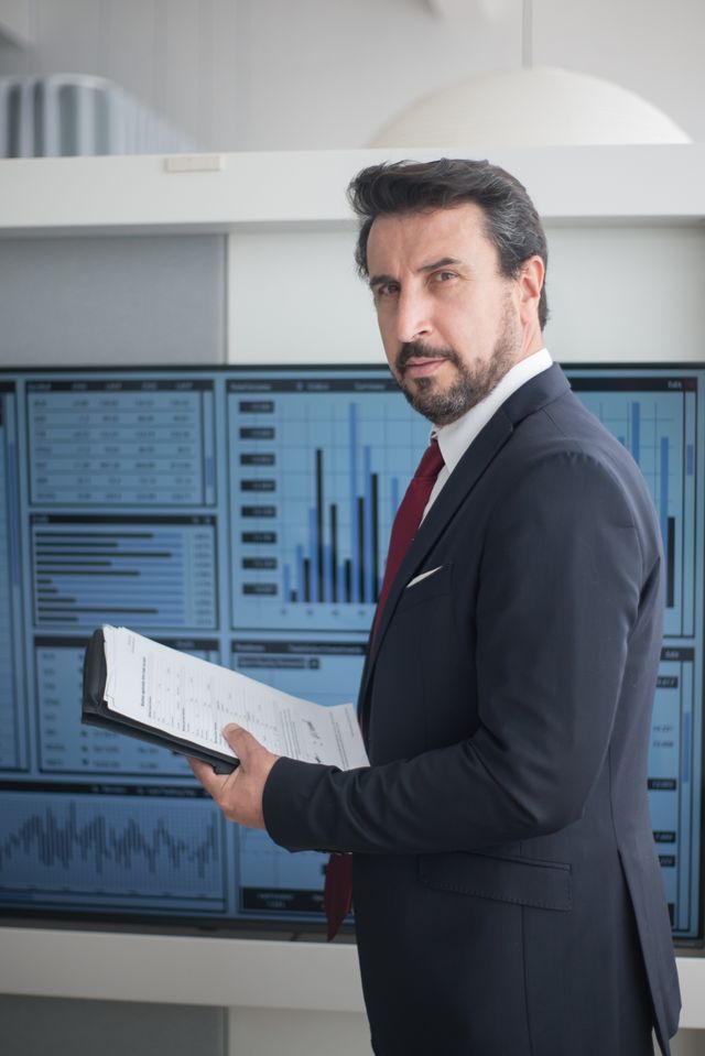 Homme en costume tenant un dossier devant des écrans affichant des graphiques financiers