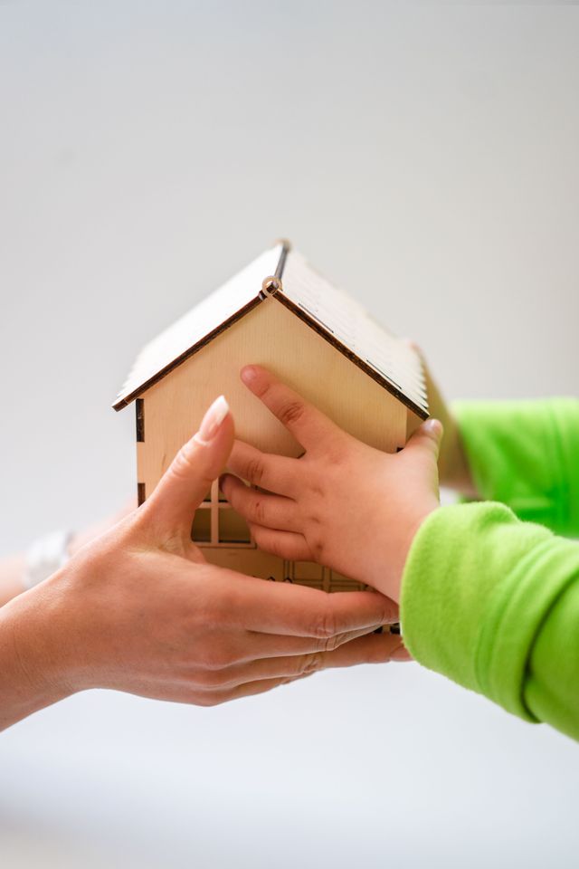 Main d'un adulte et main d'un enfant en pull vert tenant ensemble une petite maison en bois