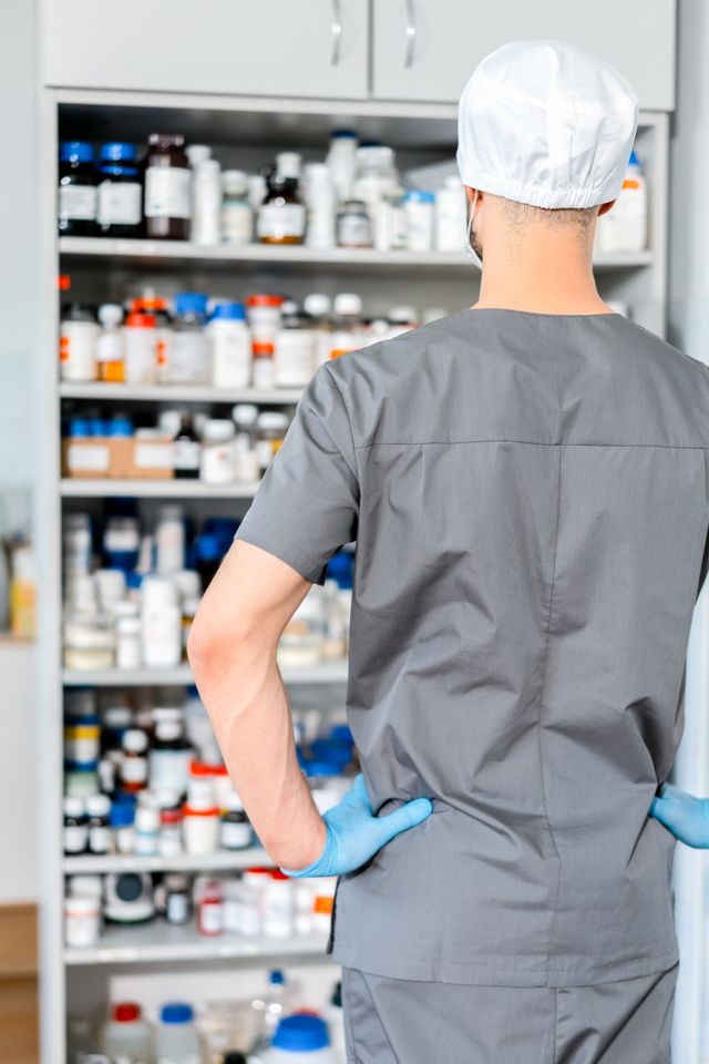 Pharmacien en tenue grise et gants bleus, de dos, face à des étagères remplies de flacons