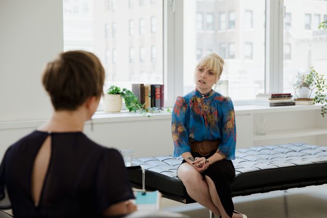 Femme assise sur un canapé échangeant avec une interlocutrice de dos dans un salon lumineux