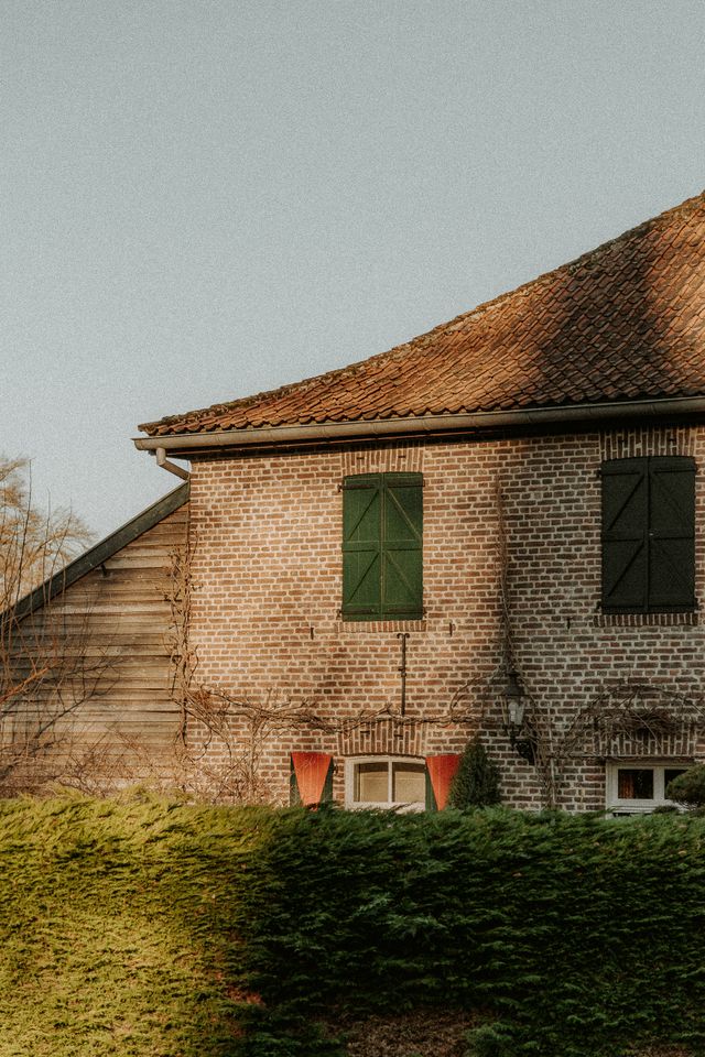 Maison ancienne en briques aux volets verts et toit de tuiles, haie taillée au premier plan