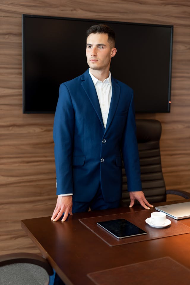 Jeune homme en costume bleu debout derrière une table de réunion en bois, écran au mur