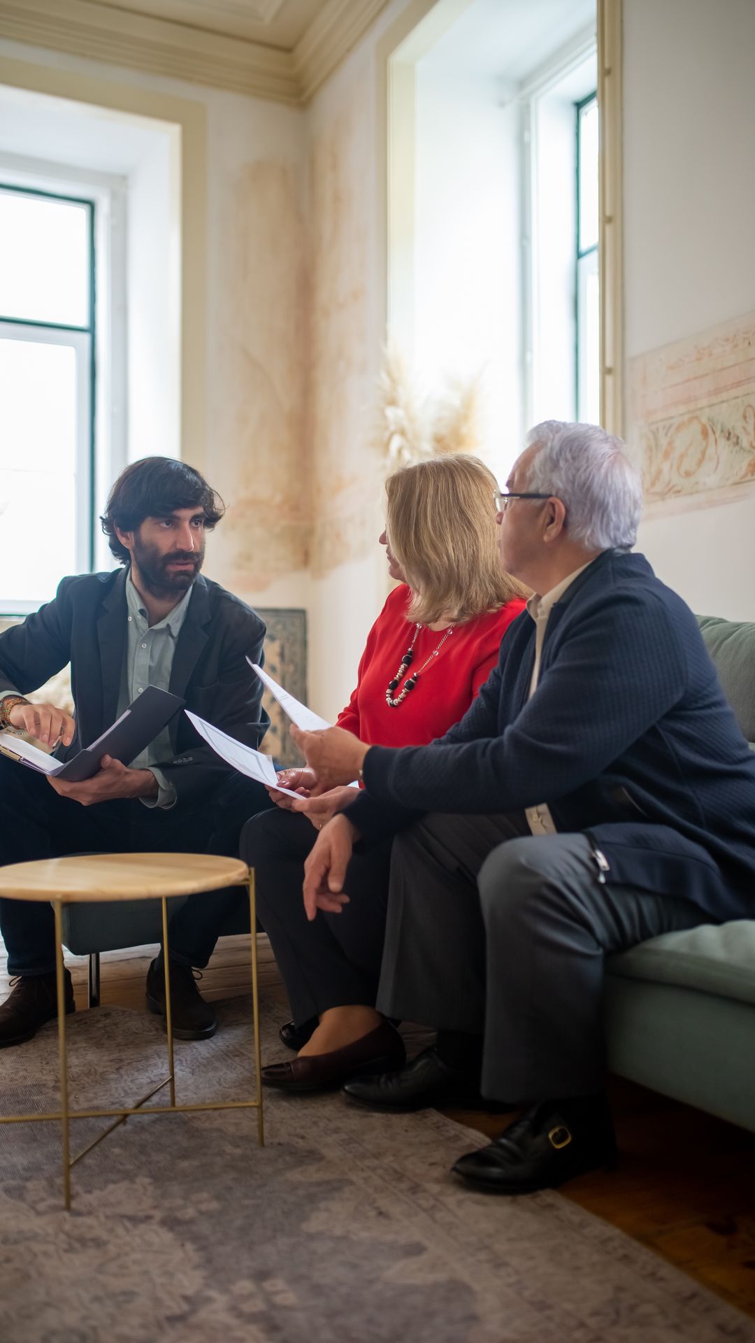Conseiller barbu échangeant avec un couple de seniors assis sur un canapé, documents en main, dans un salon clair