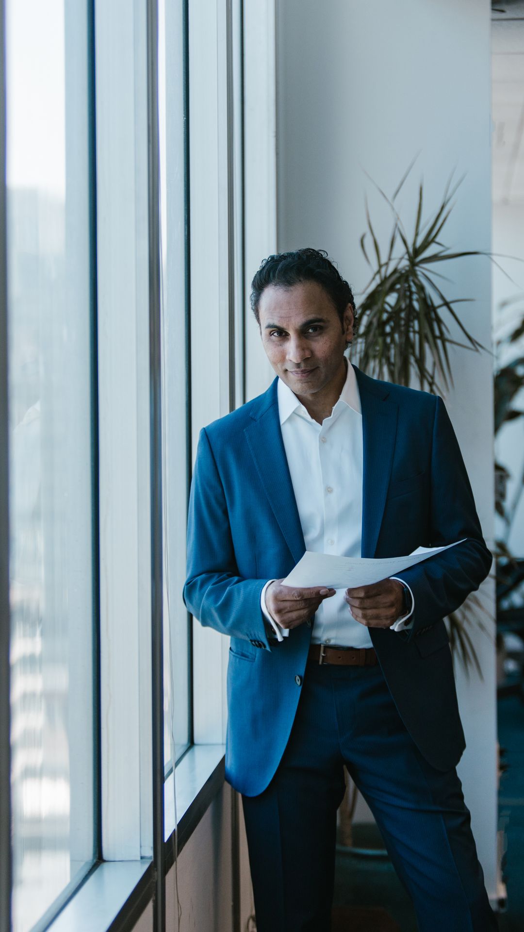 Homme en costume bleu tenant des documents, debout près d'une grande baie vitrée dans un bureau lumineux