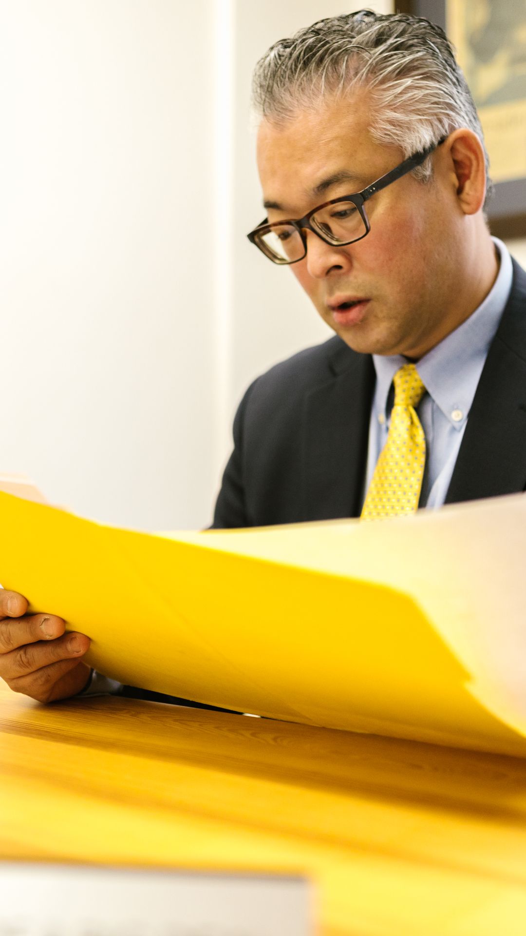 Homme aux cheveux grisonnants en costume et cravate jaune lisant attentivement un dossier ouvert à son bureau