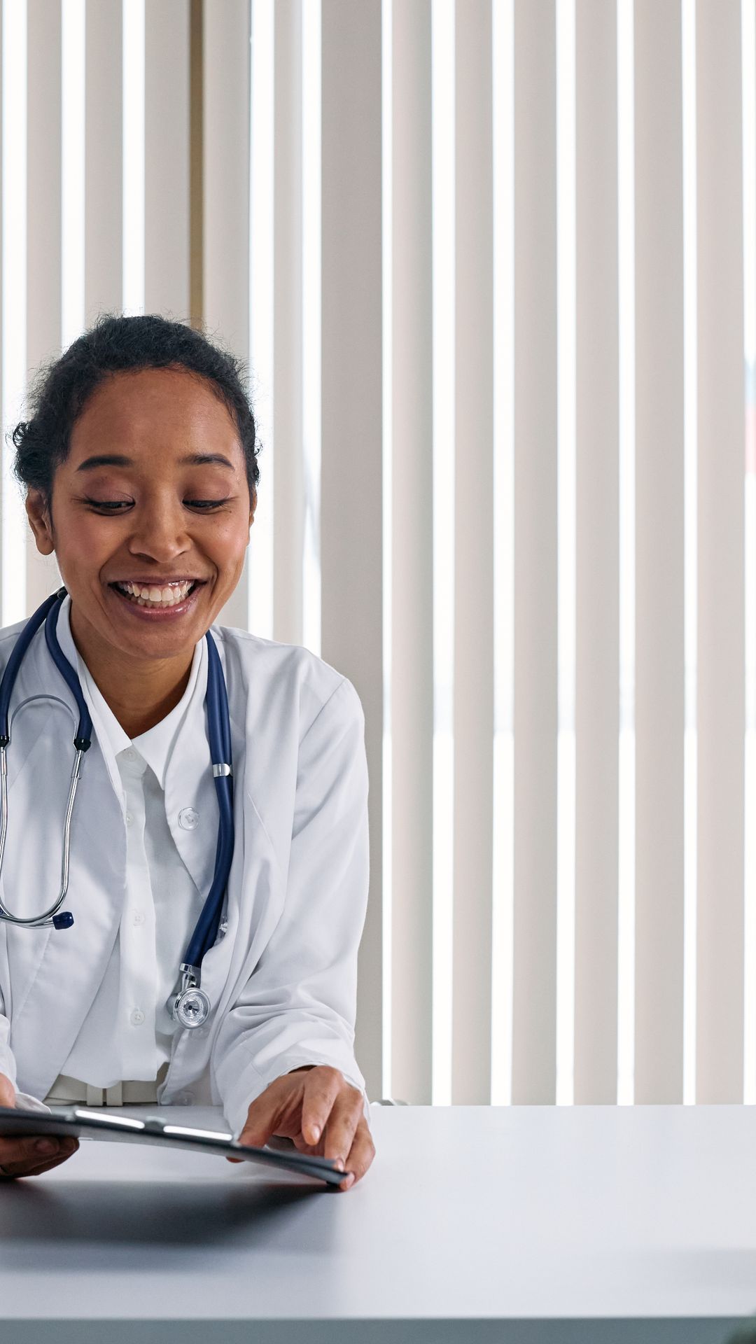Médecin souriante en blouse blanche avec stéthoscope, une tablette en main, face à un patient dans son cabinet