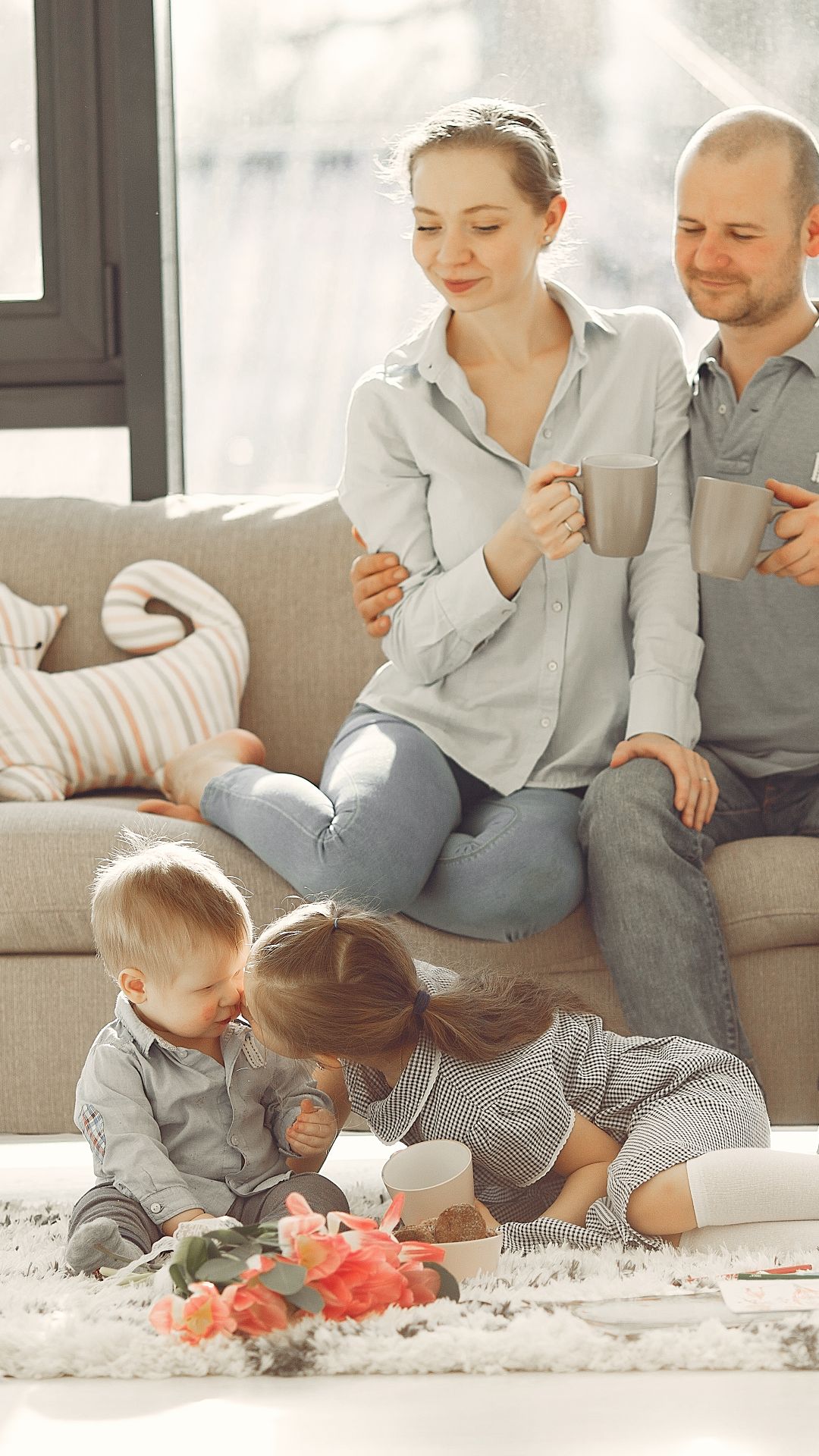 Couple assis sur un canapé, tasse à la main, regardant leurs deux jeunes enfants jouer sur le tapis du salon