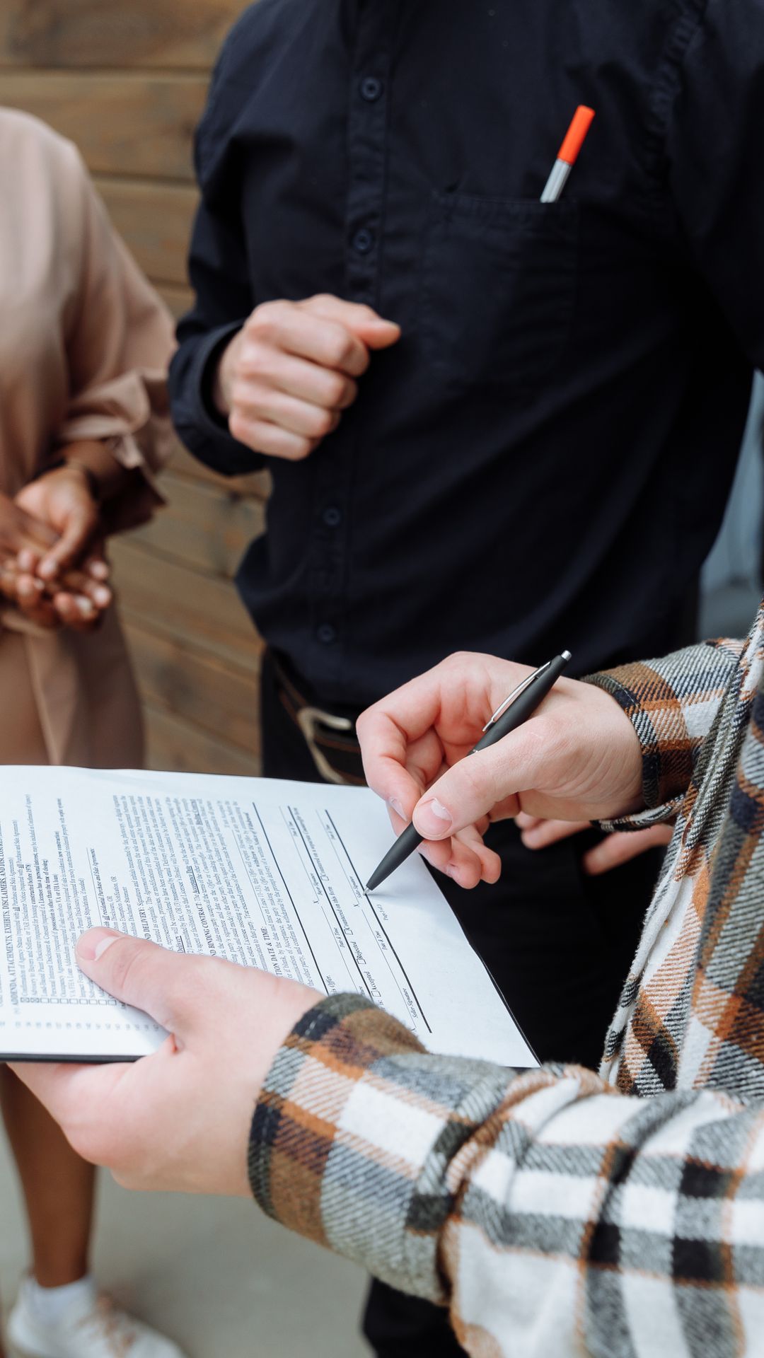 Une personne signe un document avec un stylo noir sur un porte-bloc, devant deux personnes debout