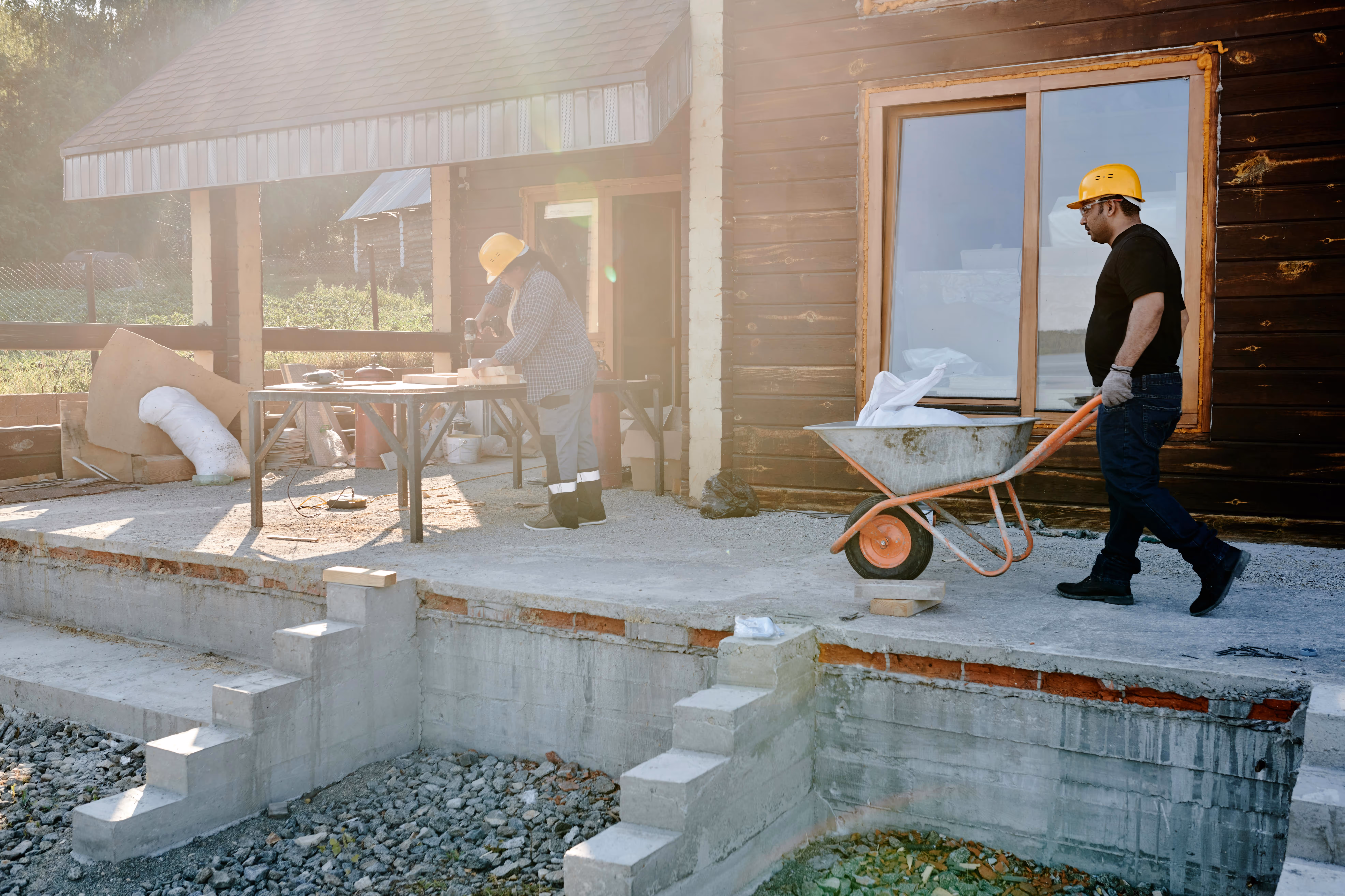 Two construction workers wearing yellow helmets at a building site, one pushing a wheelbarrow and the other working on a piece of wood on a table.
