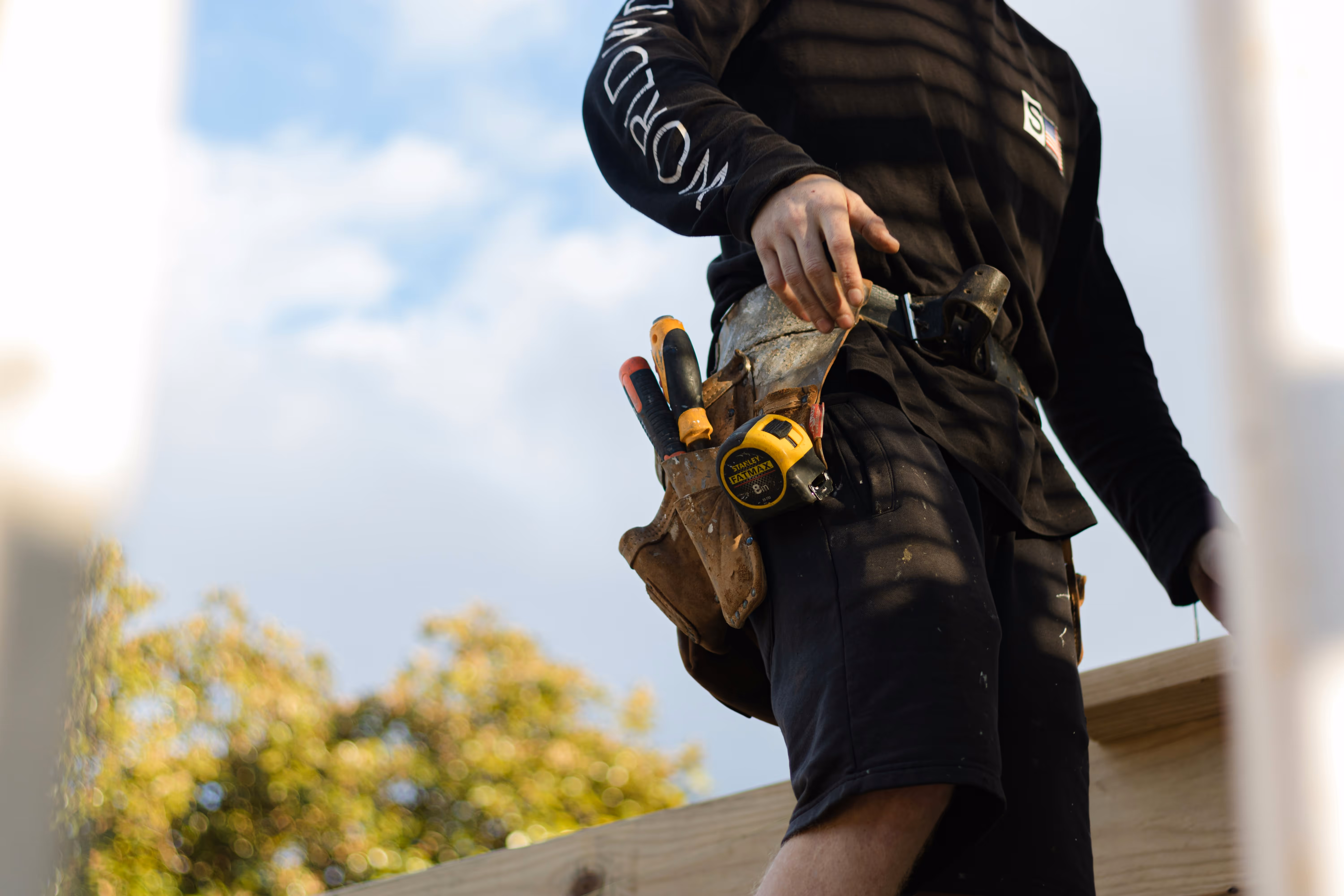 Construction worker wearing a tool belt with various hand tools and a tape measure against a blue sky background.