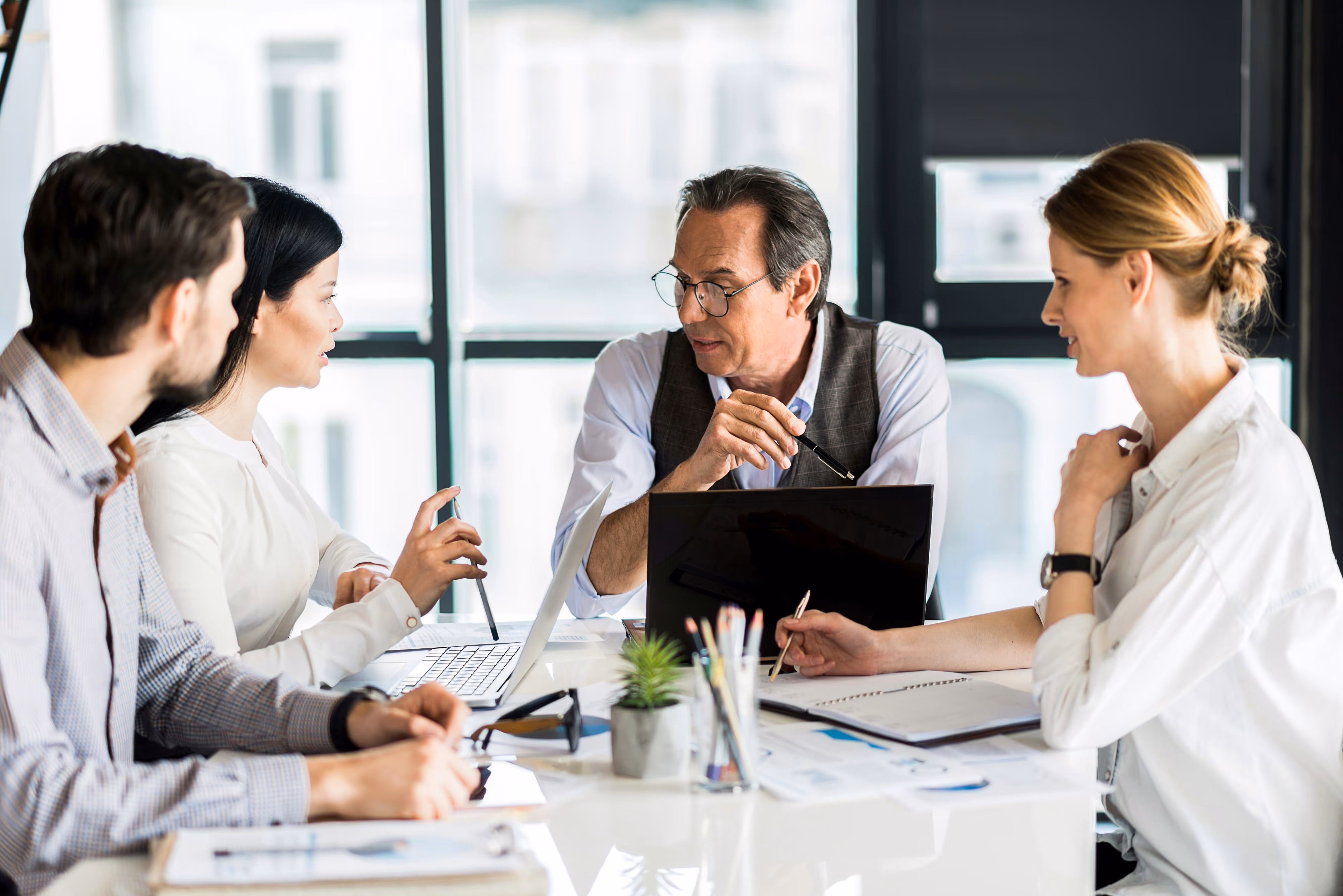 Four colleagues engaged in a work discussion around a table with laptops and documents in a bright office.