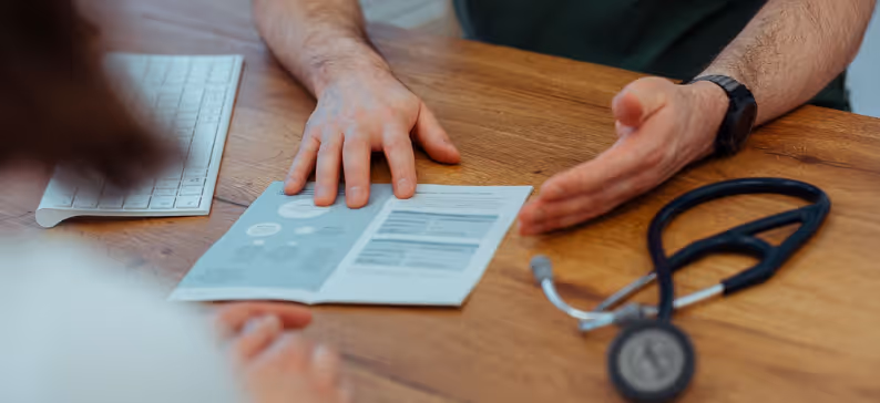 Two people sitting at a wooden desk discussing a pamphlet with a stethoscope beside them.