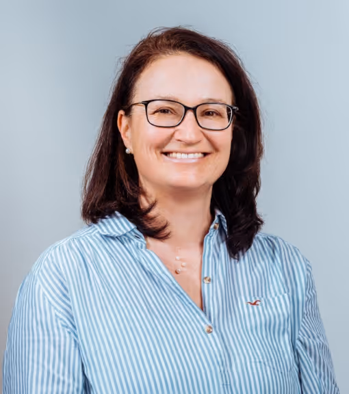 Smiling woman with glasses and shoulder-length dark hair wearing a blue and white striped shirt against a light gray background.