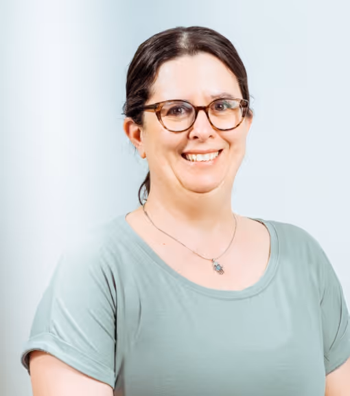 Smiling woman with glasses and dark hair wearing a light green shirt and necklace against a plain light background.