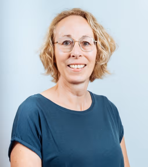 Smiling woman with short blonde hair and glasses wearing a teal shirt against a light background.