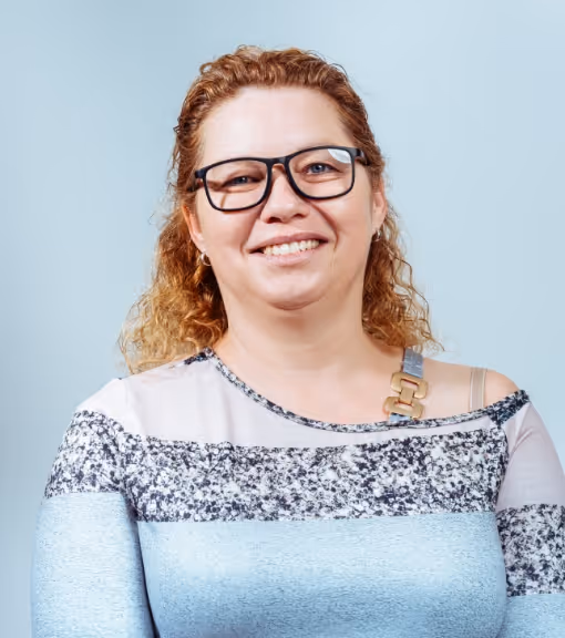 Smiling woman with curly light brown hair, wearing glasses and a patterned top with an off-shoulder strap.