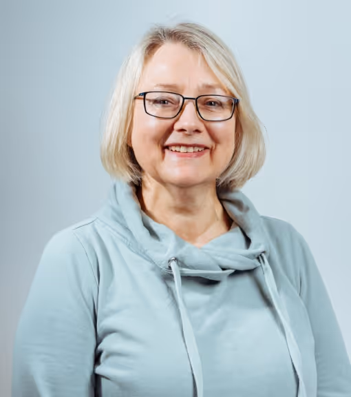 Smiling middle-aged woman with blonde hair and glasses wearing a light blue cowl-neck sweater.