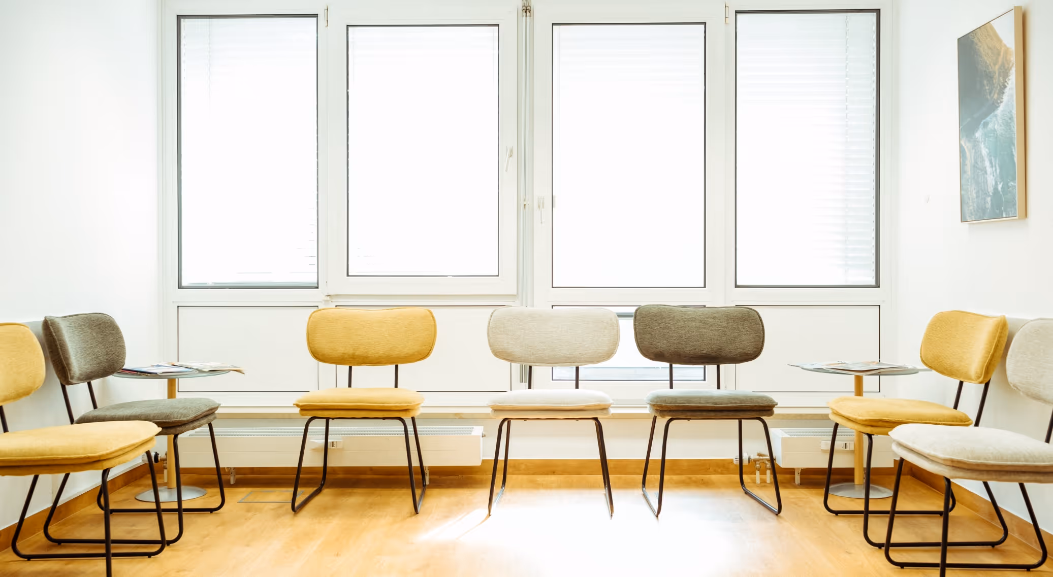 Bright waiting room with modern chairs in yellow, gray, and beige arranged along walls under large windows.