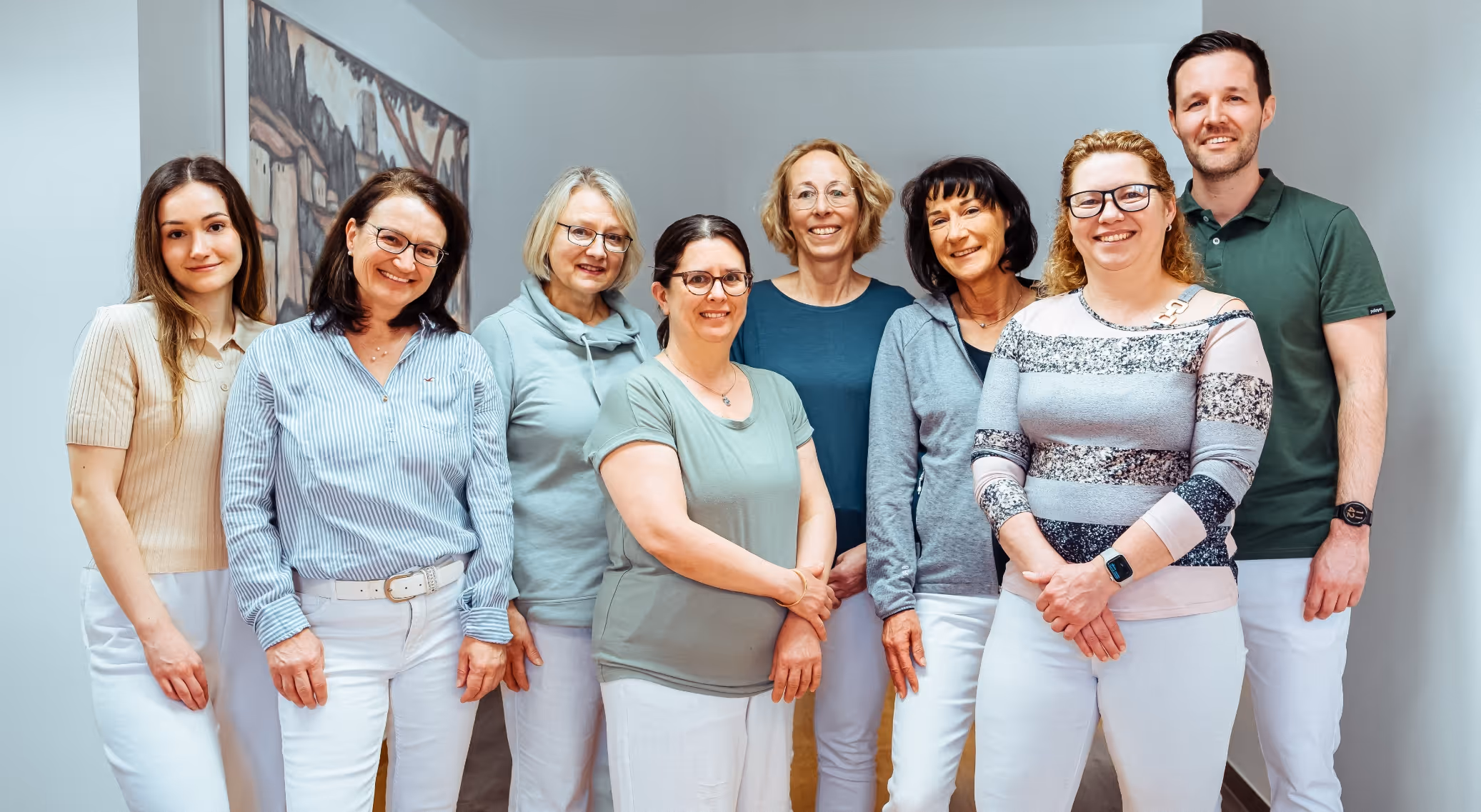 Group of eight smiling people standing indoors in casual and semi-casual attire, with a painting on the wall behind them.