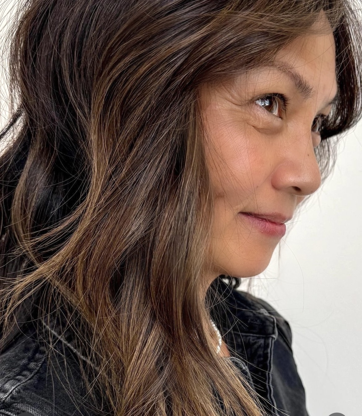 Close-up side profile of a smiling woman with long, wavy brown hair wearing a black jacket.