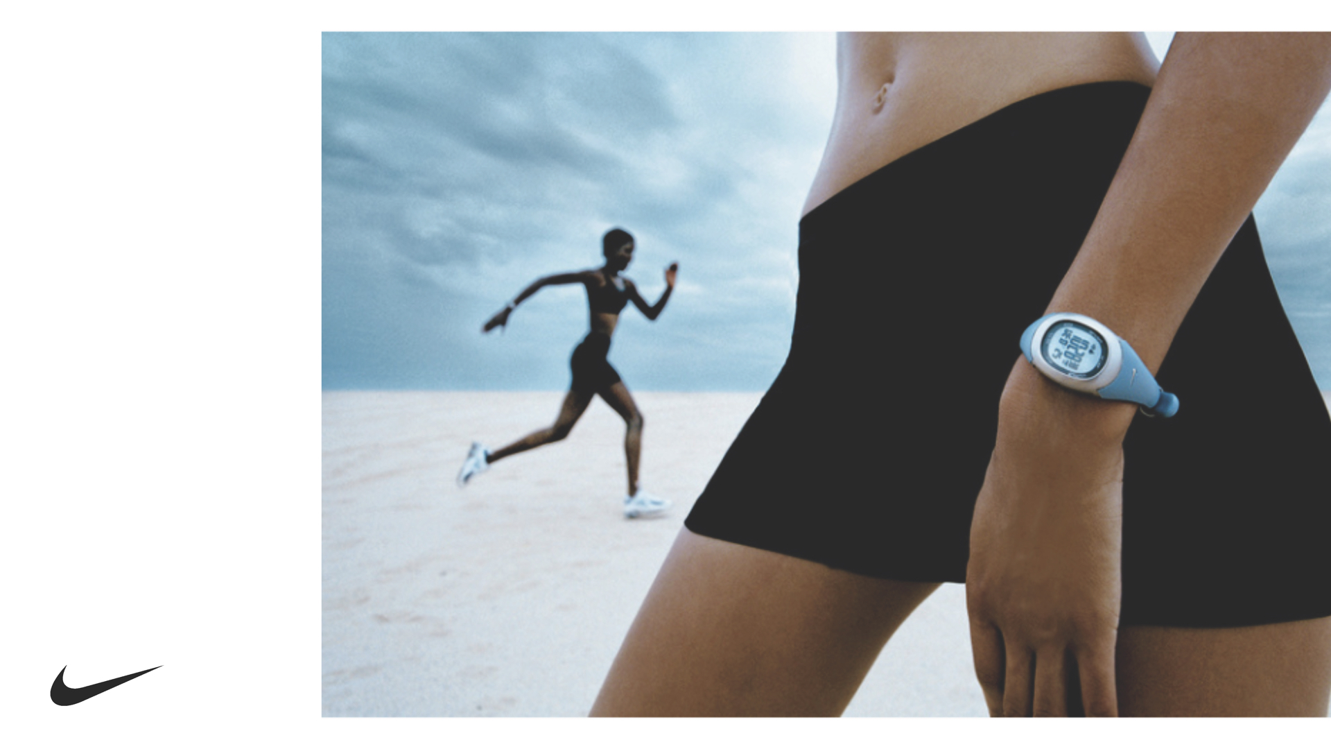 Close-up of a runner's wrist with a blue digital sports watch and a blurred runner in the background on a sandy surface under a cloudy sky.