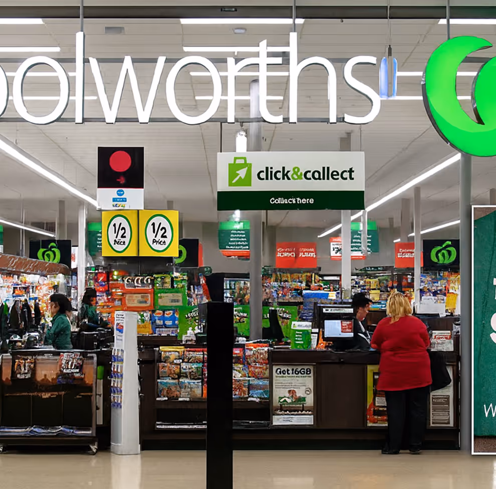 Inside a Woolworths supermarket showing checkout counters with staff assisting customers and a 'click & collect' sign overhead.