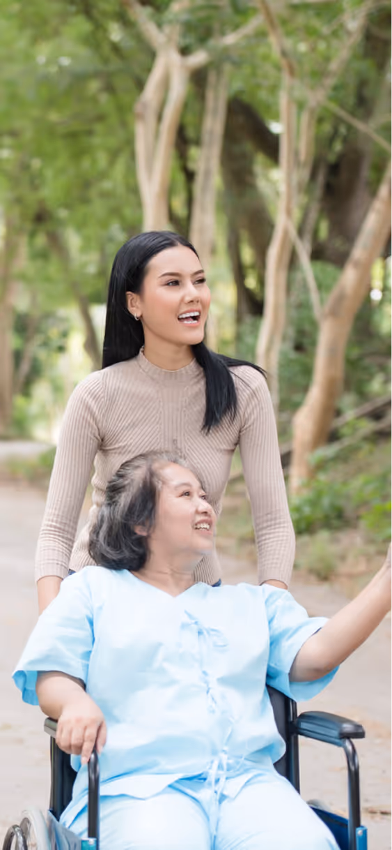 Young woman smiling and pushing an elderly woman in a wheelchair outdoors on a tree-lined path.