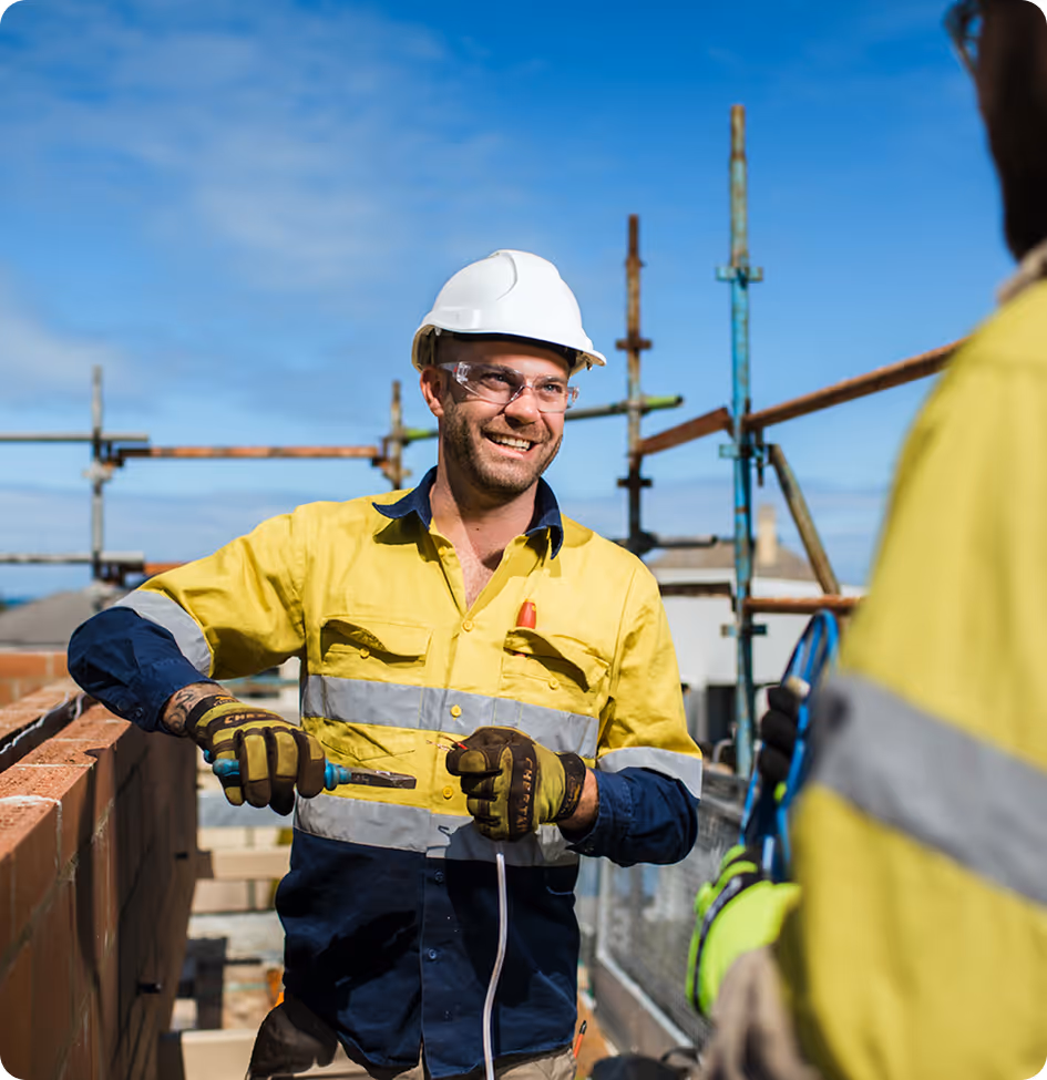Smiling construction worker wearing a white hard hat and yellow safety shirt works with a tool on wiring at a construction site.