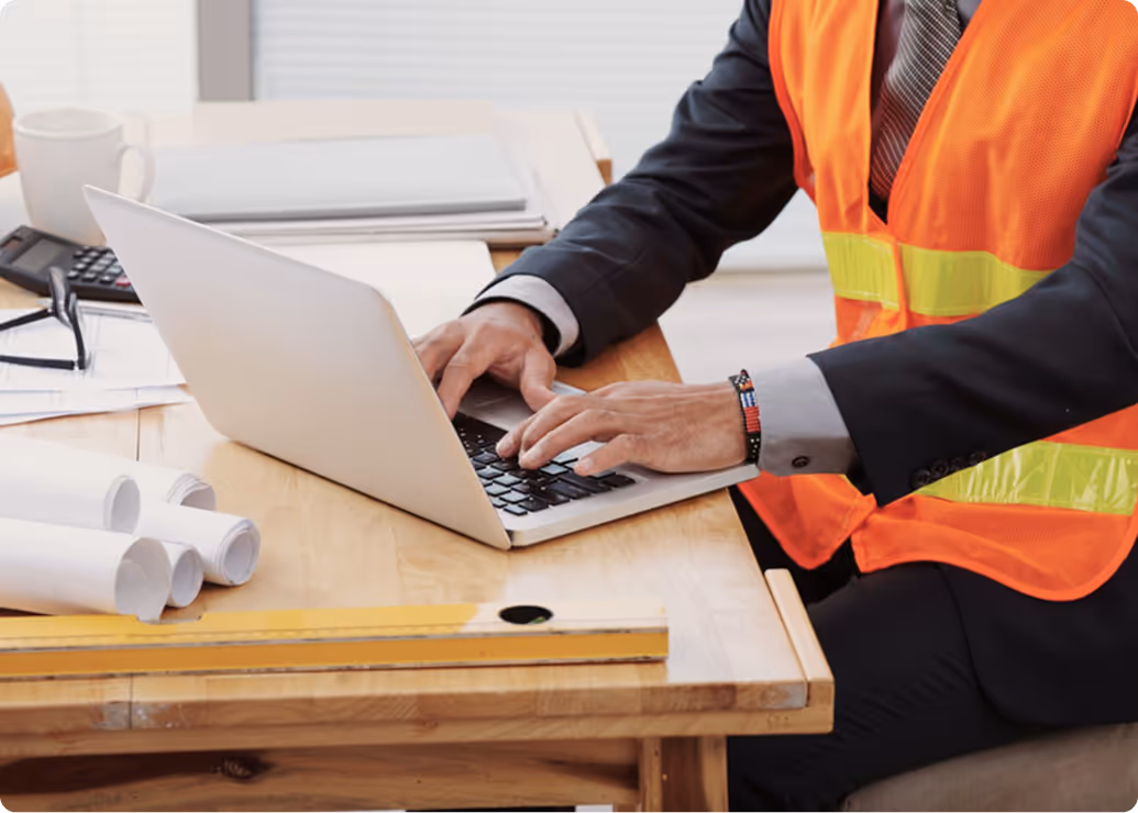 Person wearing an orange safety vest typing on a laptop at a wooden desk with rolled-up blueprints and a measuring level.