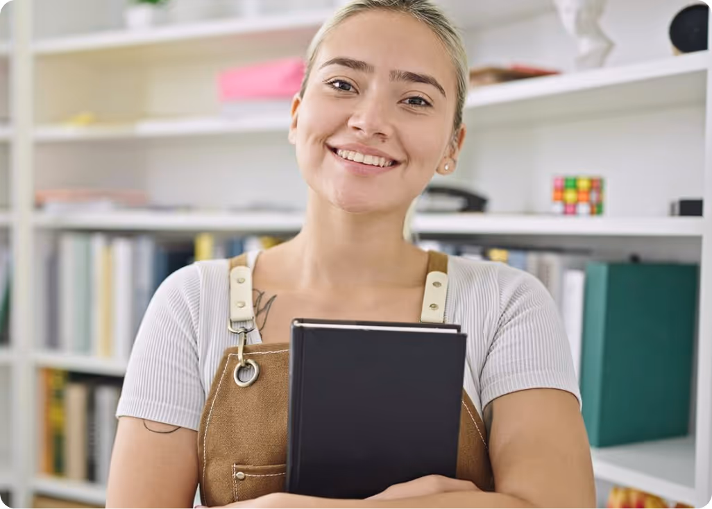 Smiling young woman with tattoos holding a closed black book in front of a bookshelf.