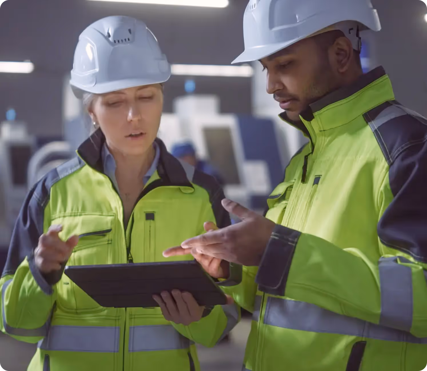 Two industrial workers wearing white helmets and neon yellow safety jackets discussing data on a tablet in a factory setting.