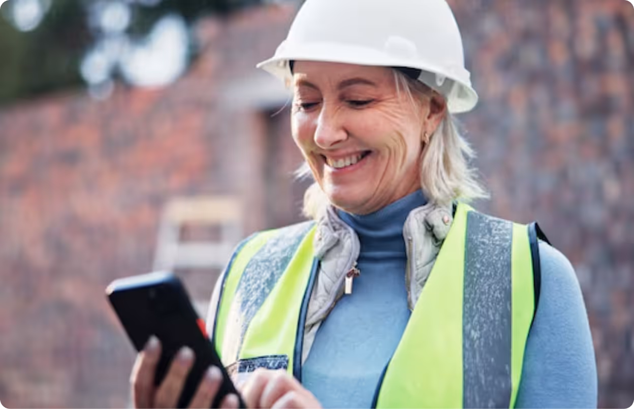 Smiling female construction worker wearing a white hard hat and reflective vest, looking at a smartphone.