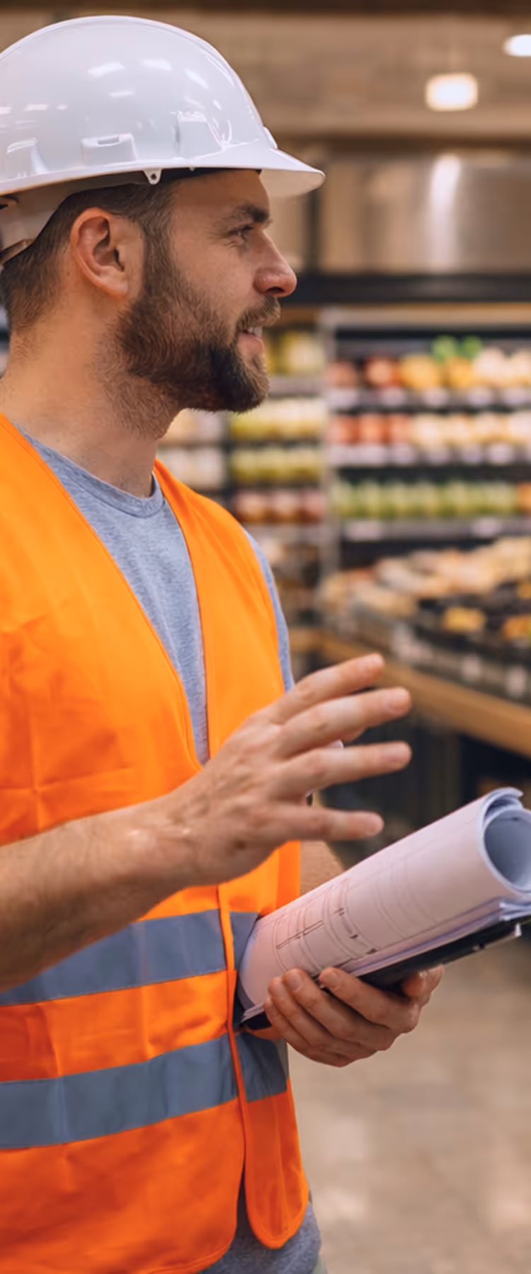 Two men, one in a hard hat and orange safety vest holding rolled blueprints, and the other in a blue shirt with a clipboard, talking in a grocery store produce aisle.