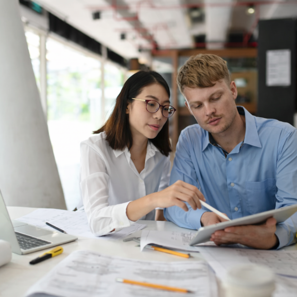 Two architects or engineers reviewing plans and discussing a project together at a desk with blueprints and a laptop.
