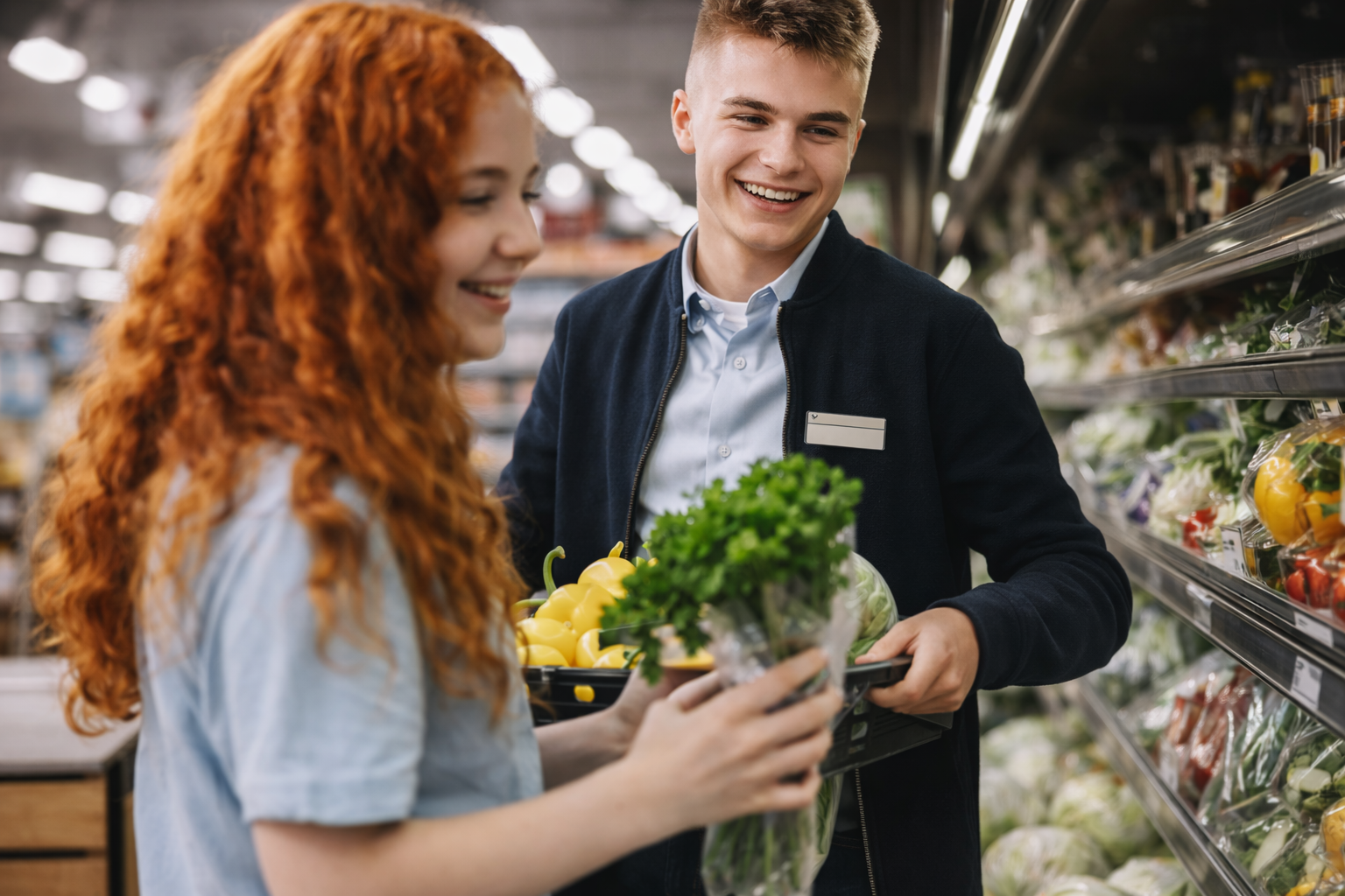 Smiling grocery clerk helping a female customer pick fresh herbs in a store produce aisle.