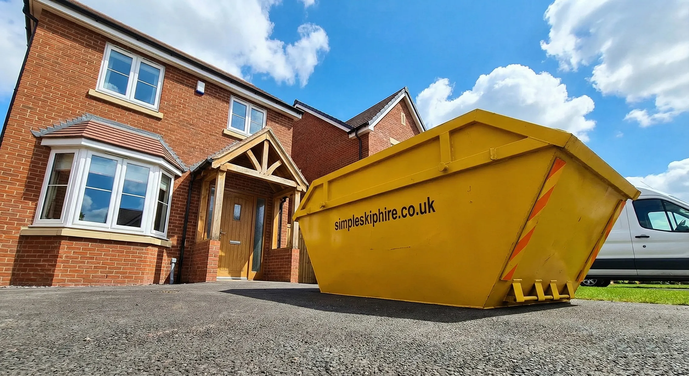 Large yellow skip bin with simpleskiphire.co.uk text parked on a driveway beside a brick house under a blue sky with clouds.