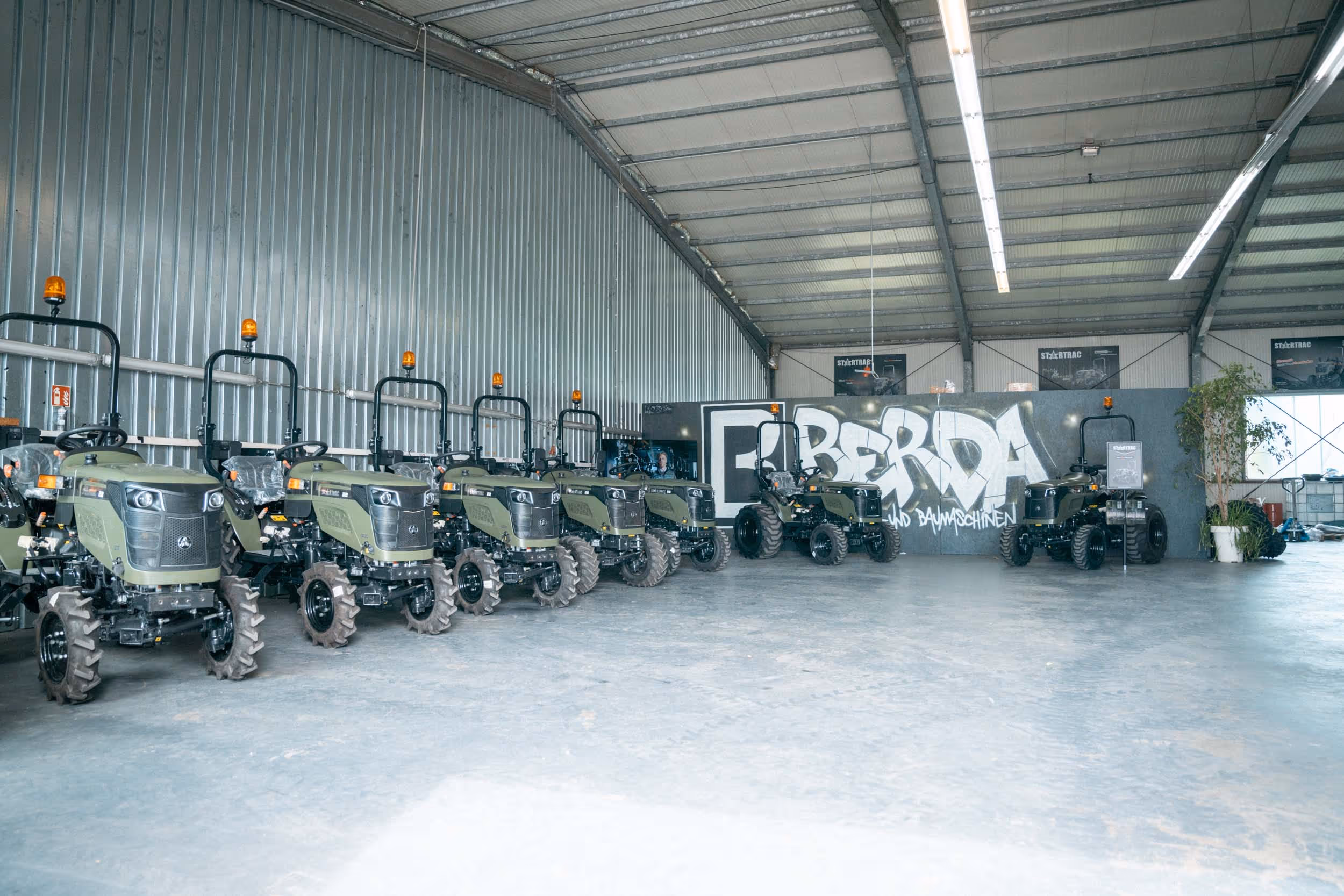 Mehrere grüne Gartentraktoren in einer großen Lagerhalle vor einer Wand mit Graffiti 'BERDA'.