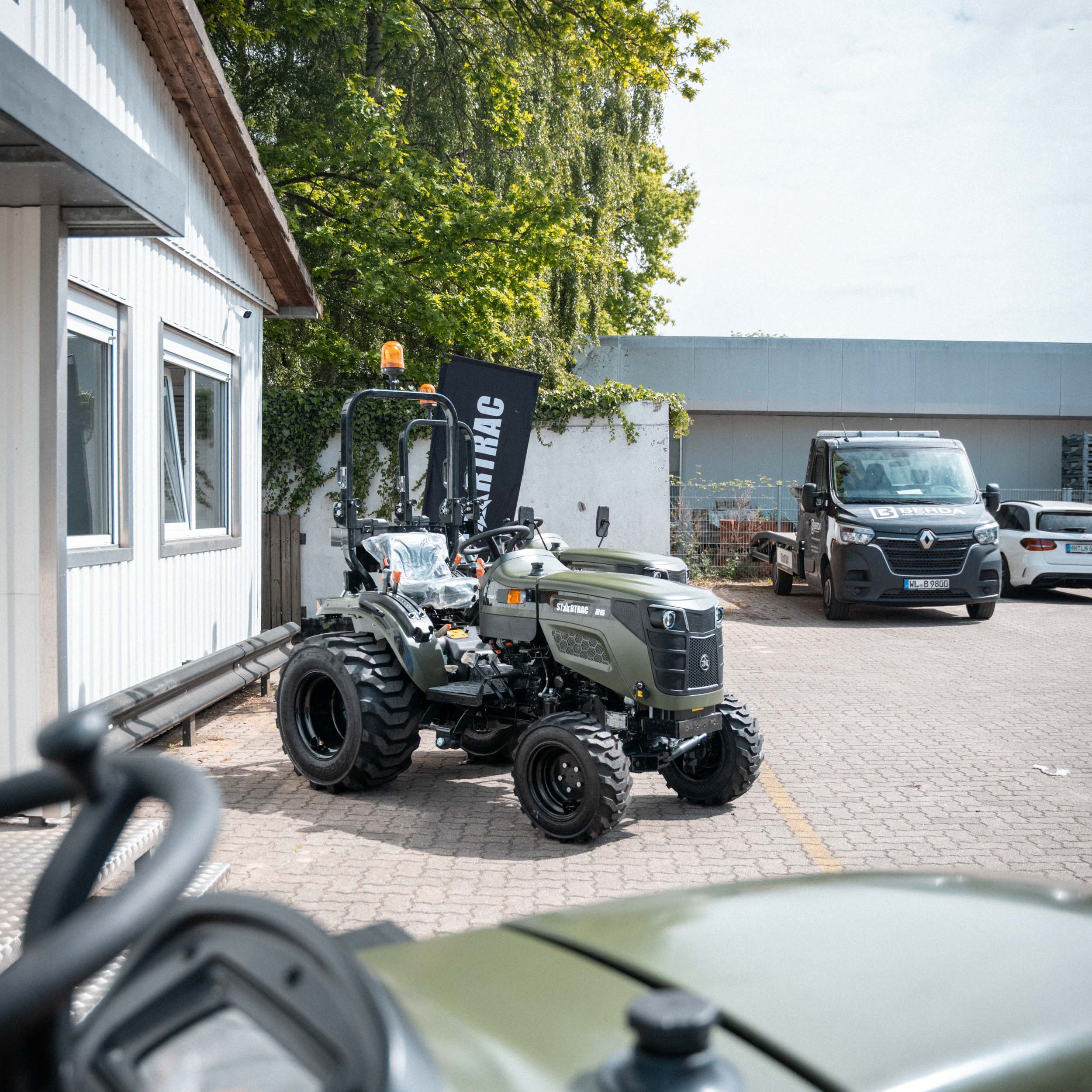 Grüner Startrac Traktor steht auf gepflastertem Hof neben einem weißen Gebäude, im Hintergrund ein schwarzes Berda-Fahrzeug und Bäume.