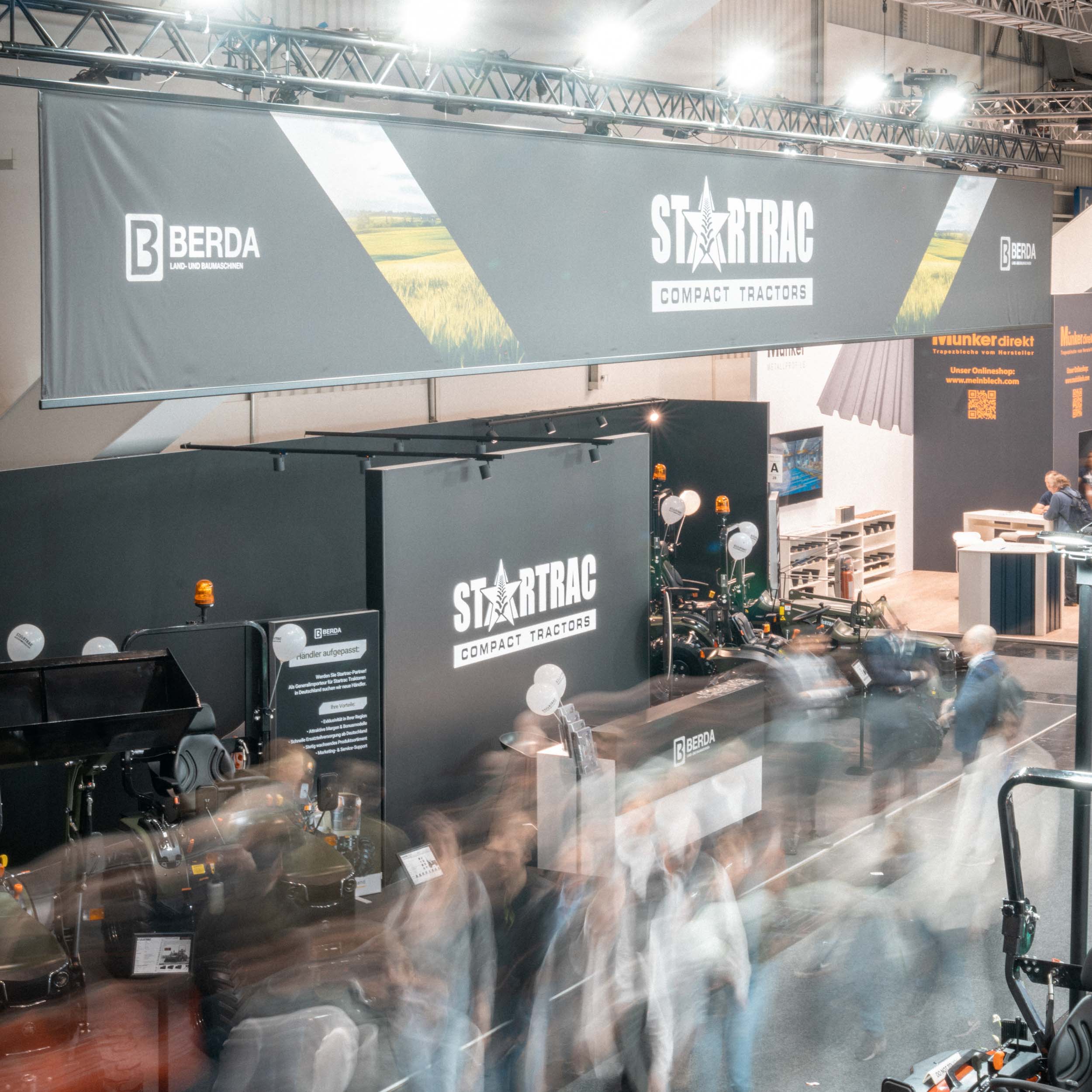 Messehalle mit dem Berda-Stand, der Startrac-Kompacktschlepper präsentiert, und verschwommene Personen in Bewegung.