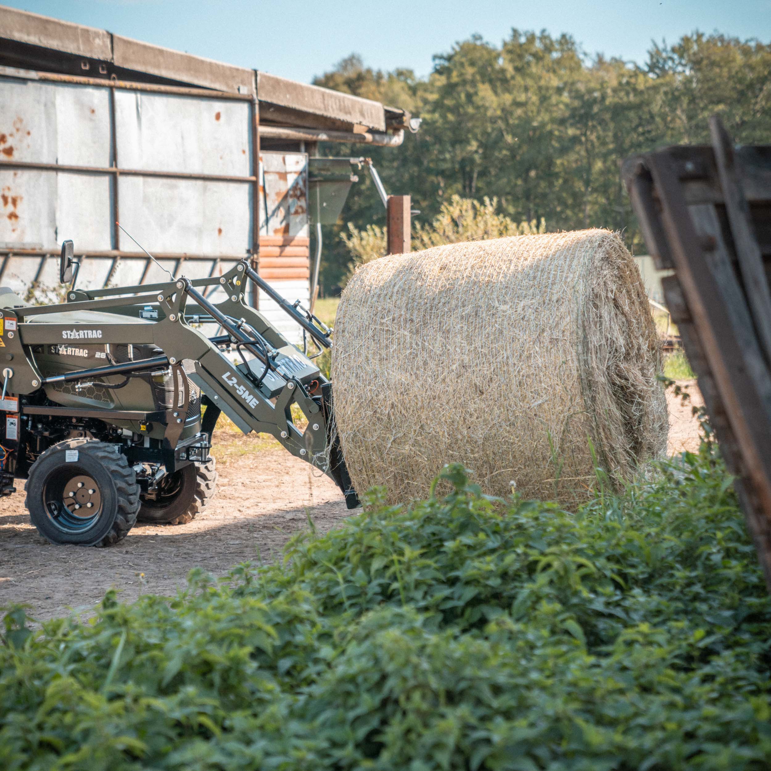 Traktor mit Frontlader hebt eine große Heuballenrolle auf einem Bauernhof.