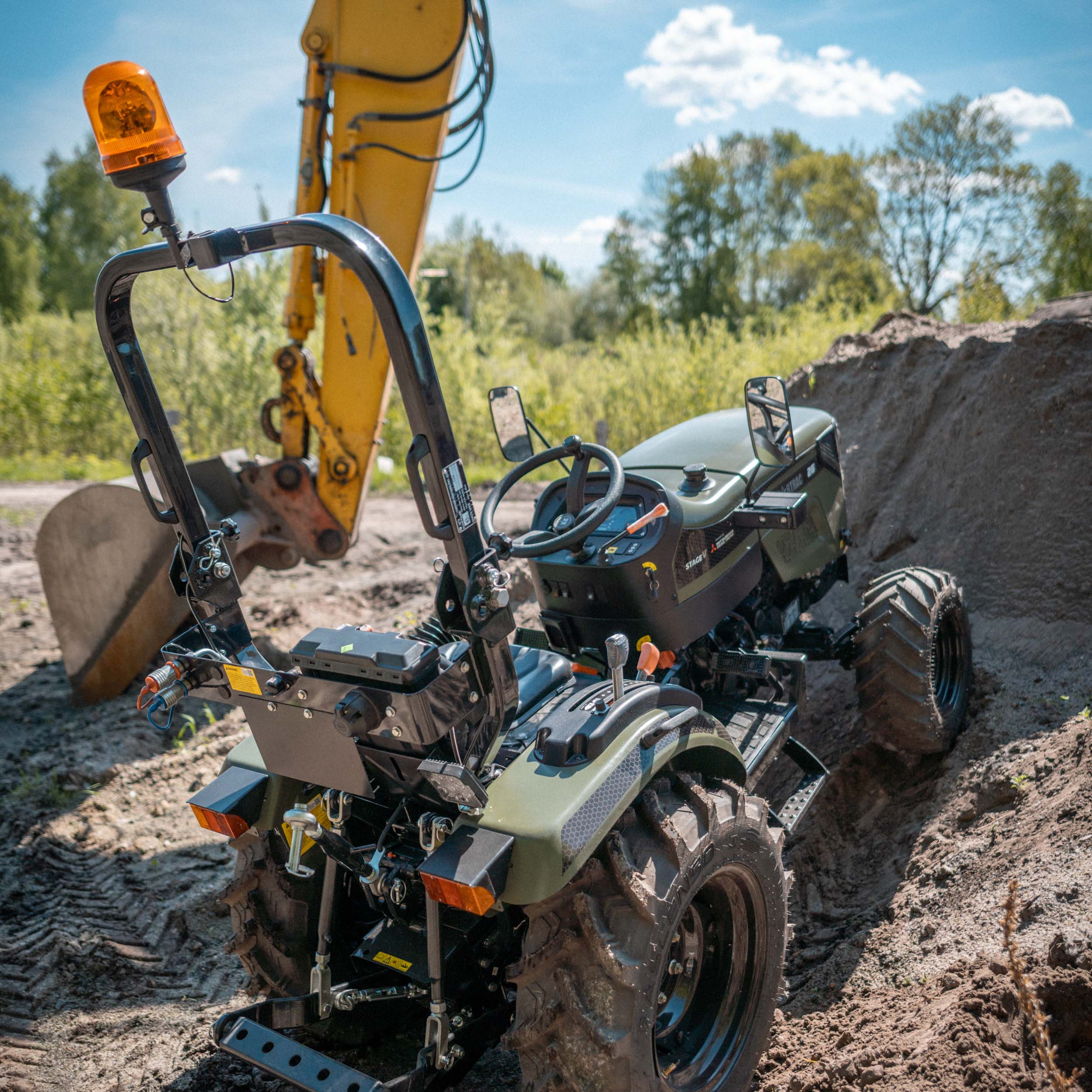 Kleiner grüner Traktor mit Rückfahrlicht steht vor einem gelben Bagger auf einer Baustelle mit Erde.