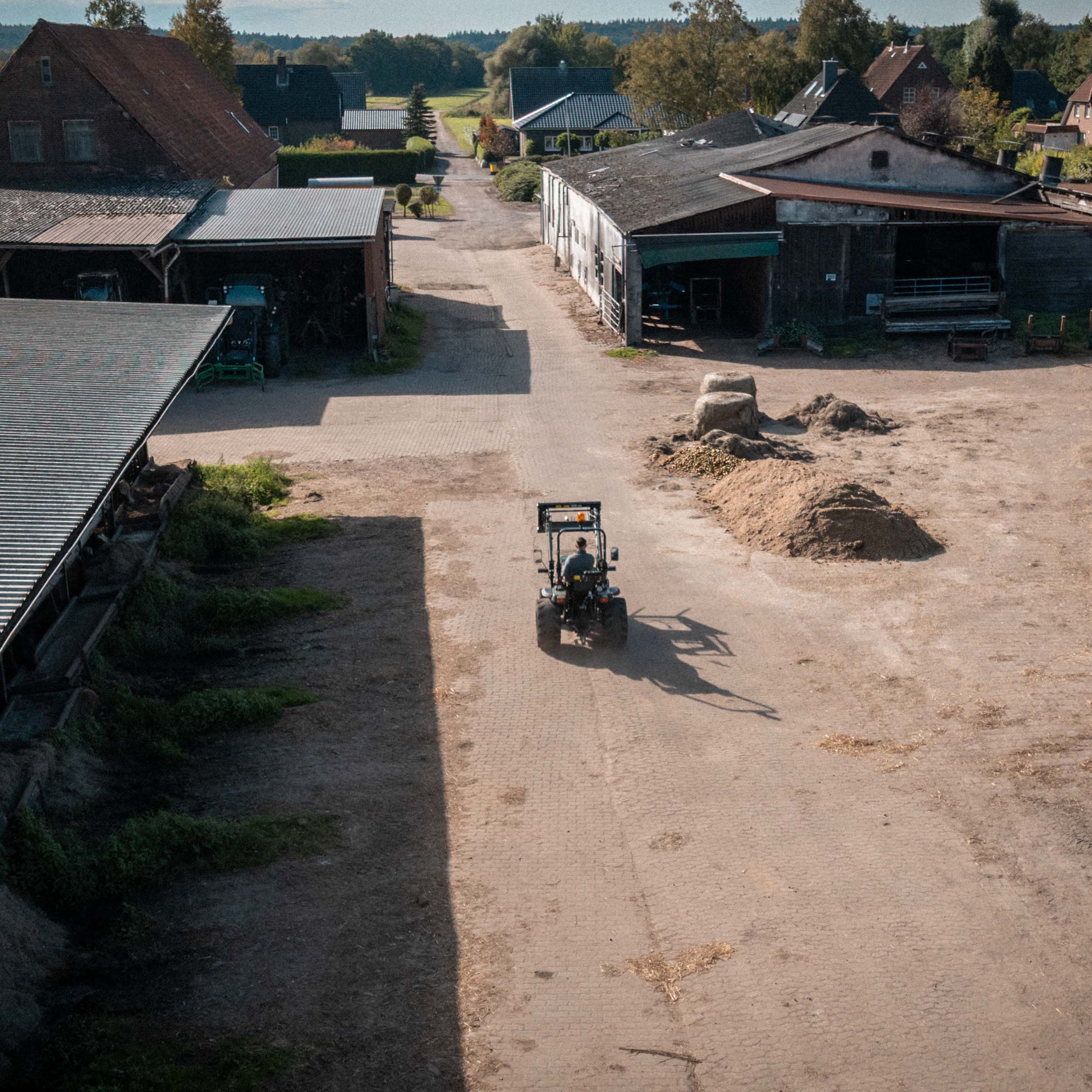 Traktor fährt auf einem Hof mit Scheunen und Hügeln aus Erde an sonnigem Tag.