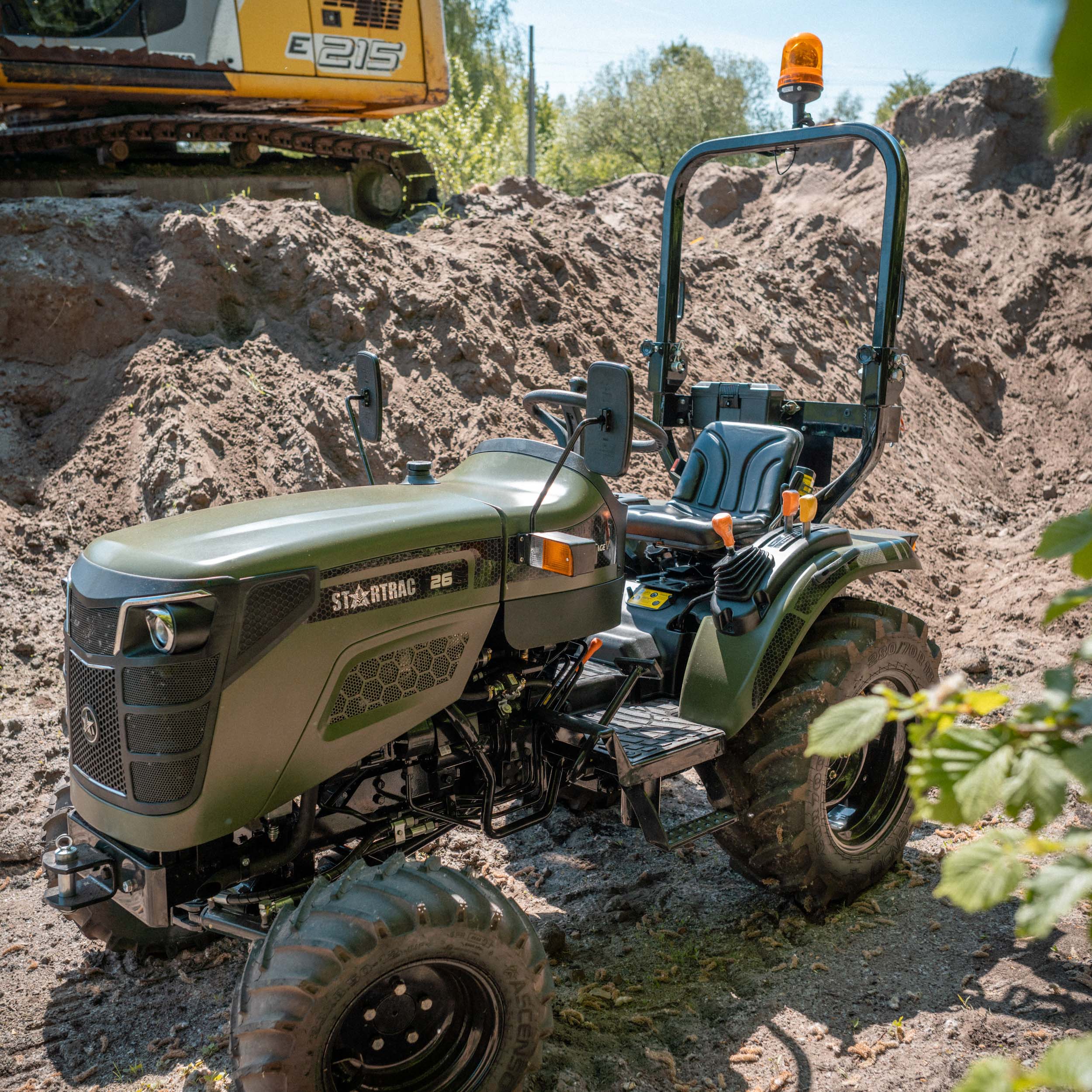 Grüner StarTrac 26 Kleintraktor mit orangefarbenem Warnlicht, geparkt vor einem Erdhügel und einem gelben Bagger.