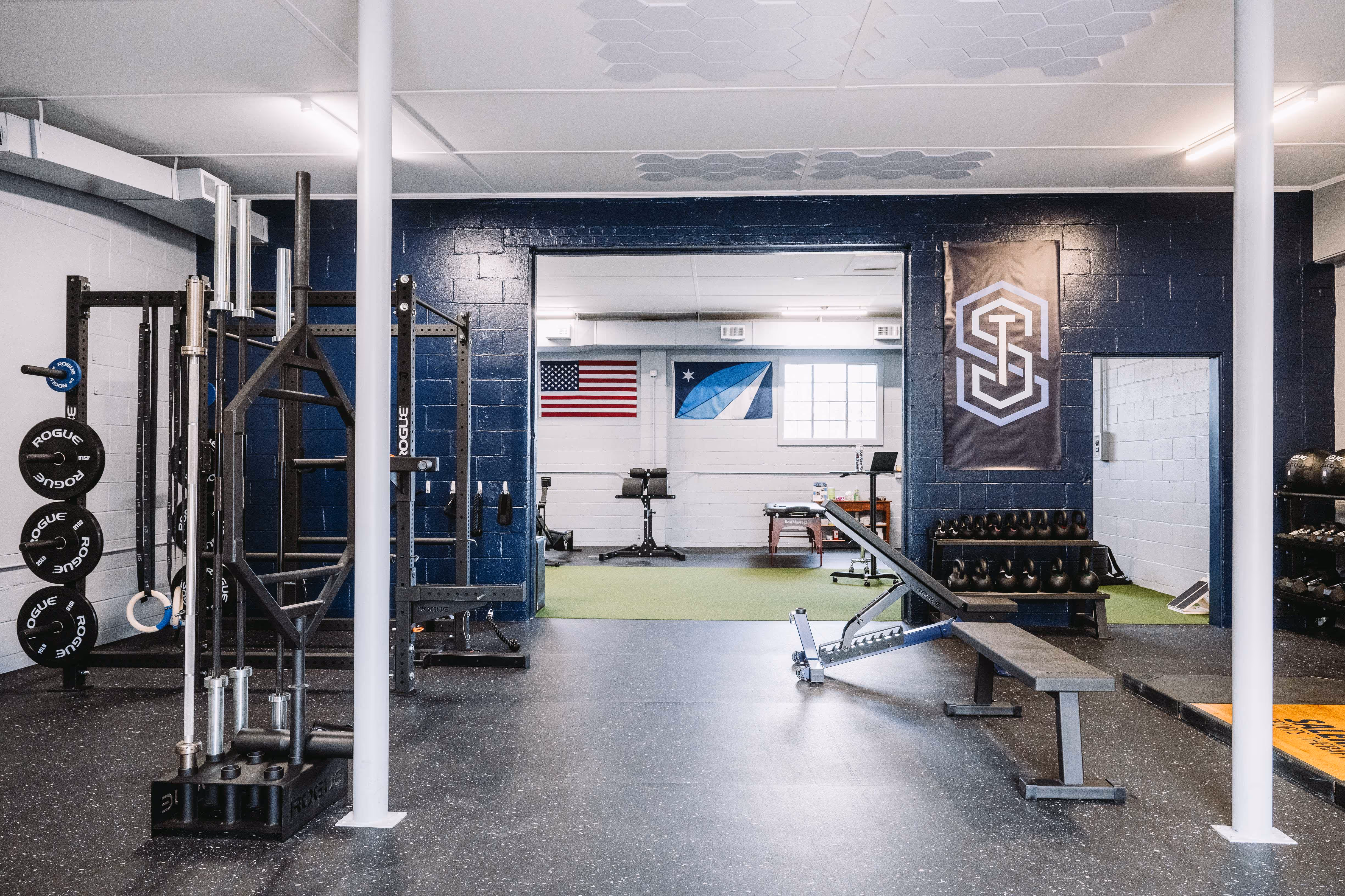 Modern gym interior with weightlifting equipment, adjustable bench, kettlebells, and flags on the back wall.