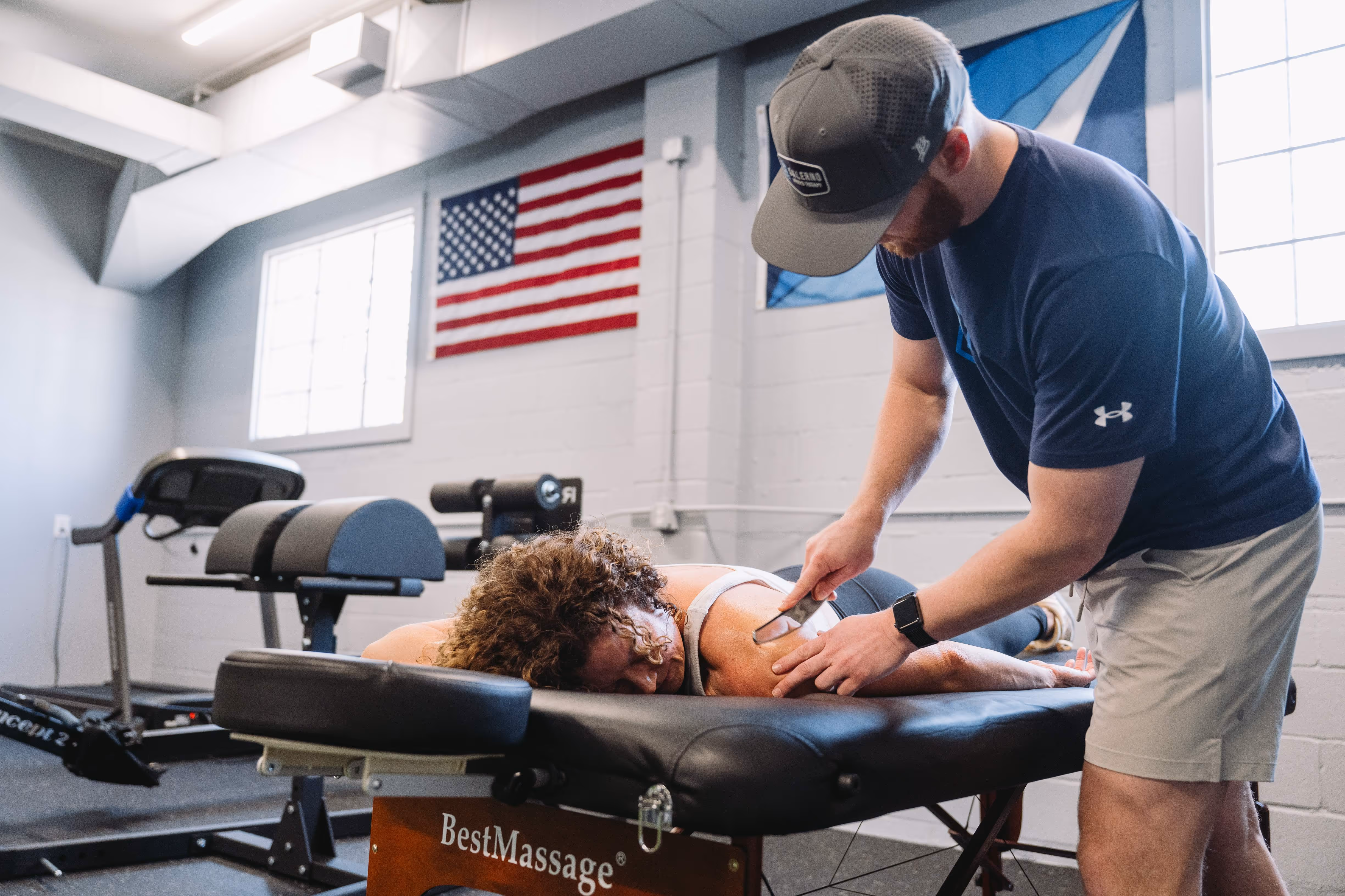 A man wearing a cap and navy shirt performs a shoulder massage on a woman lying face down on a massage table in a gym setting.