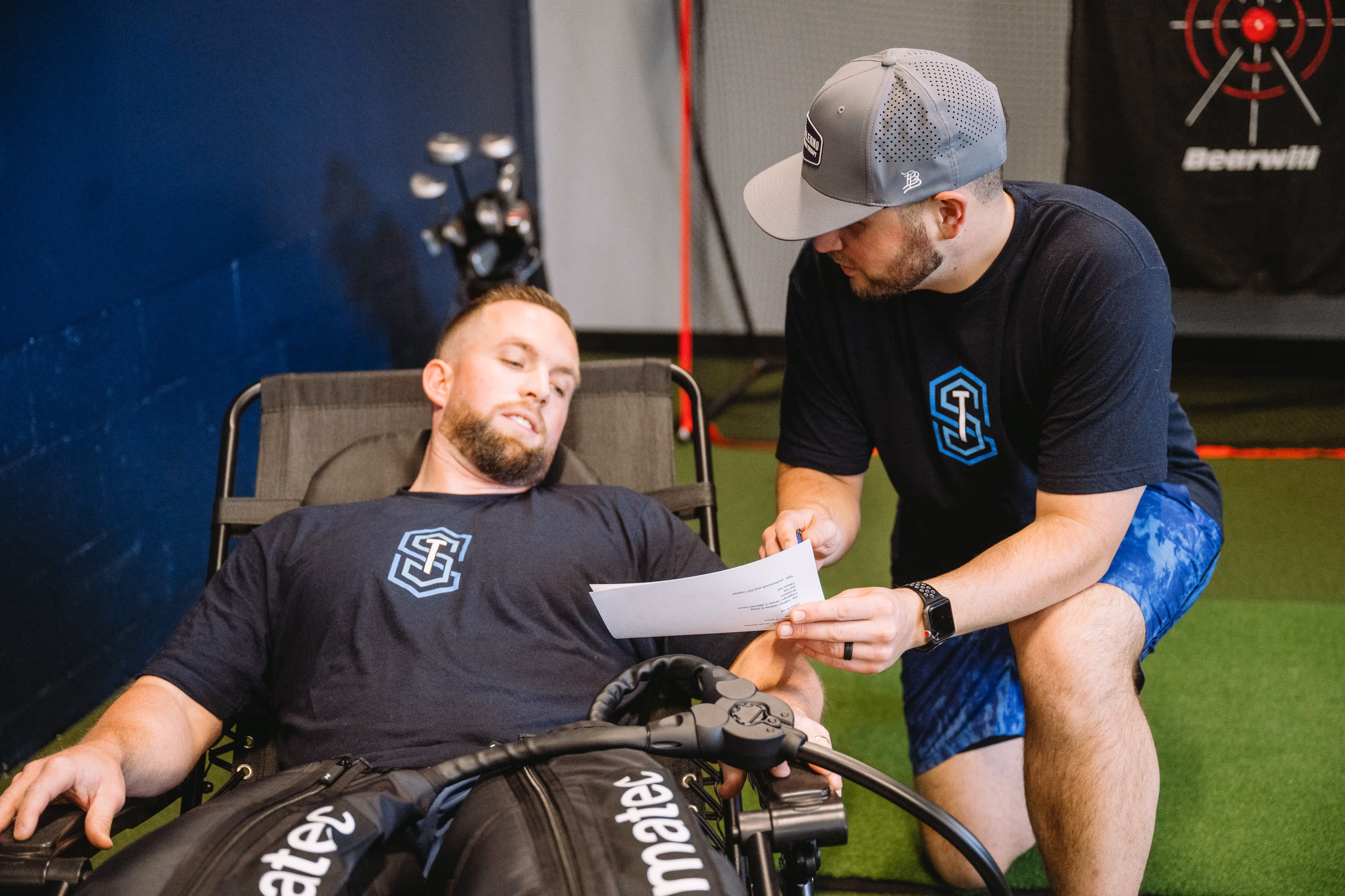 A trainer in a gray cap and blue shorts shows a paper to a man reclining on a chair wearing compression therapy boots.