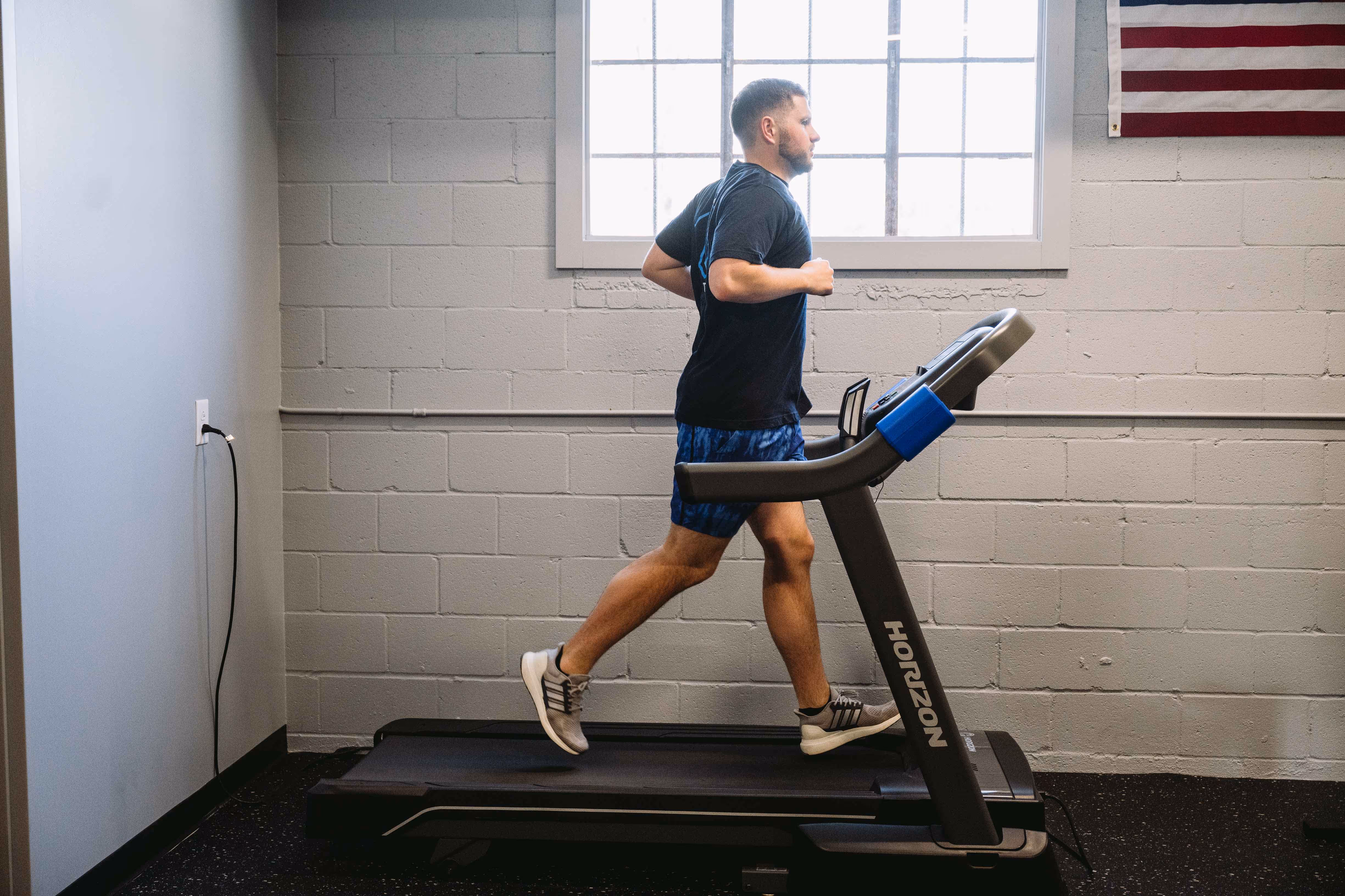 Man jogging on a treadmill in a gym room with gray brick walls and an American flag.